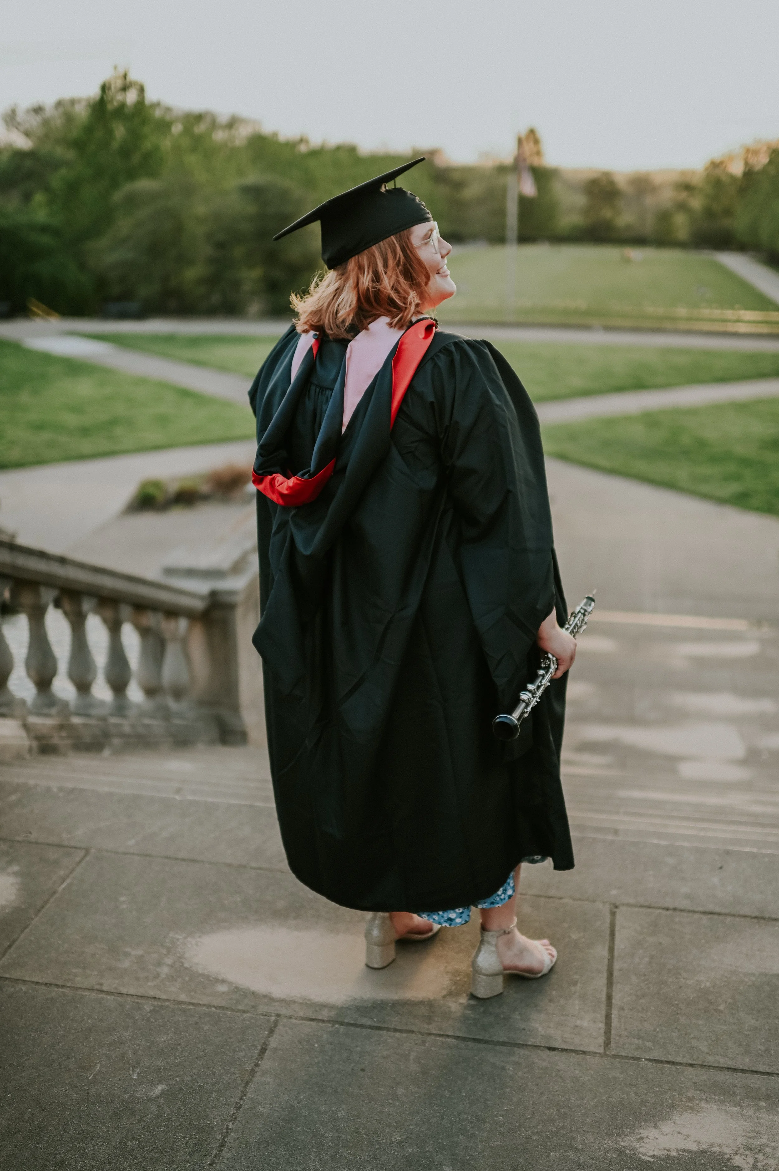 A woman in a graduation cap and gown holding a flute, standing outdoors on steps with a grassy area and trees in the background, wearing high heels and floral shorts visible below the gown.