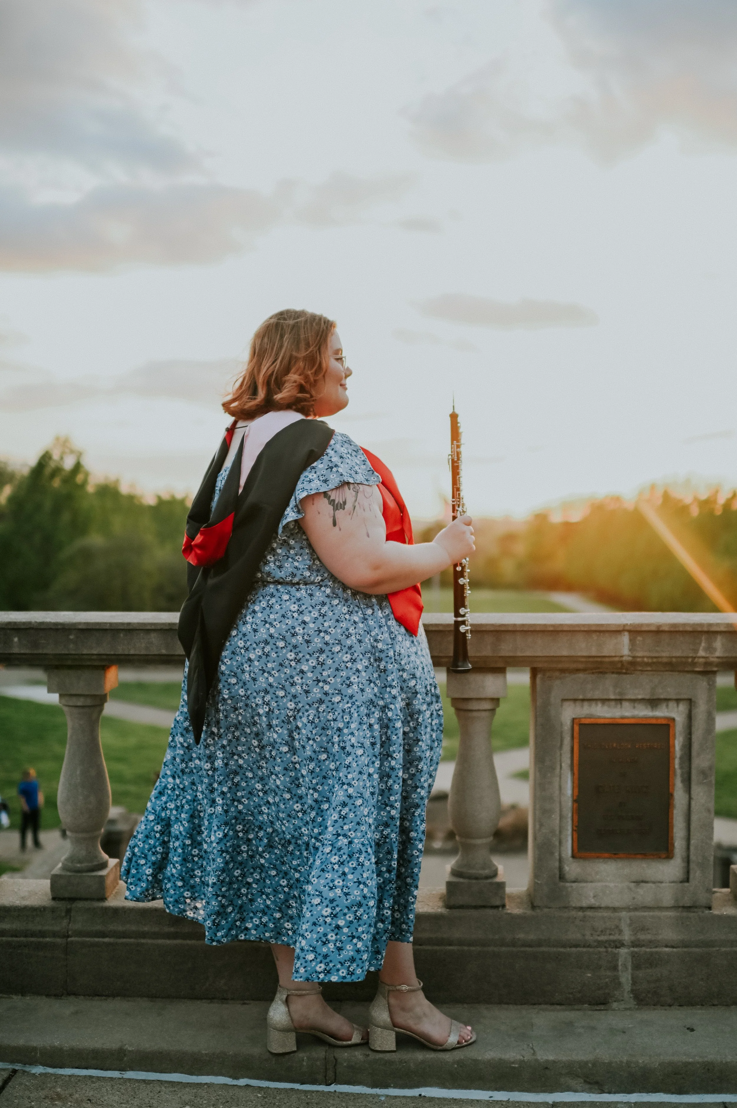 A woman with wavy, shoulder-length hair wearing glasses, a floral blue dress, and beige high-heeled shoes, holding an oboe, standing on a stone bridge at sunset.
