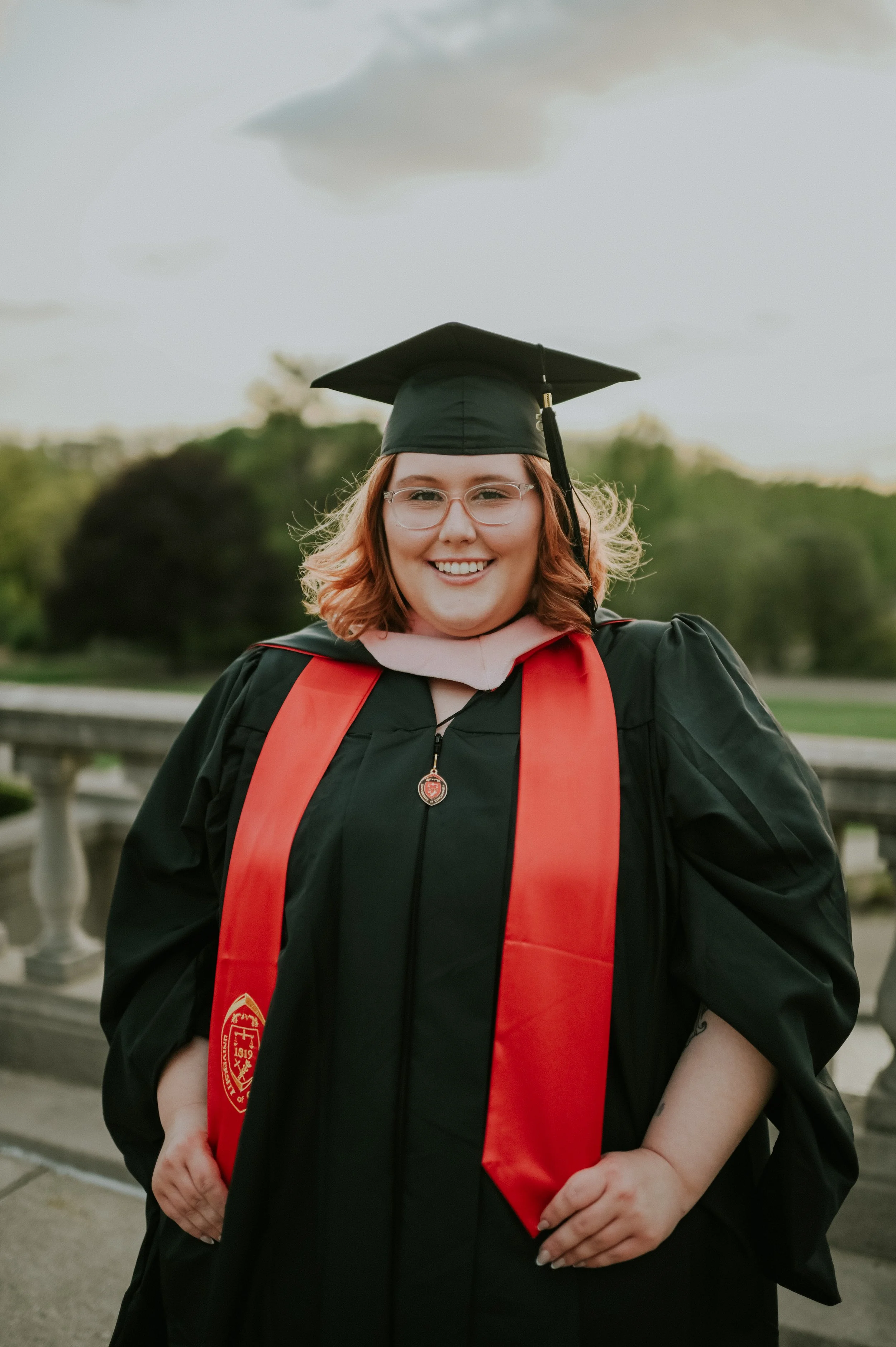 A young woman wearing a graduation cap and gown with a red stole is smiling outdoors, with trees and a cloudy sky in the background.