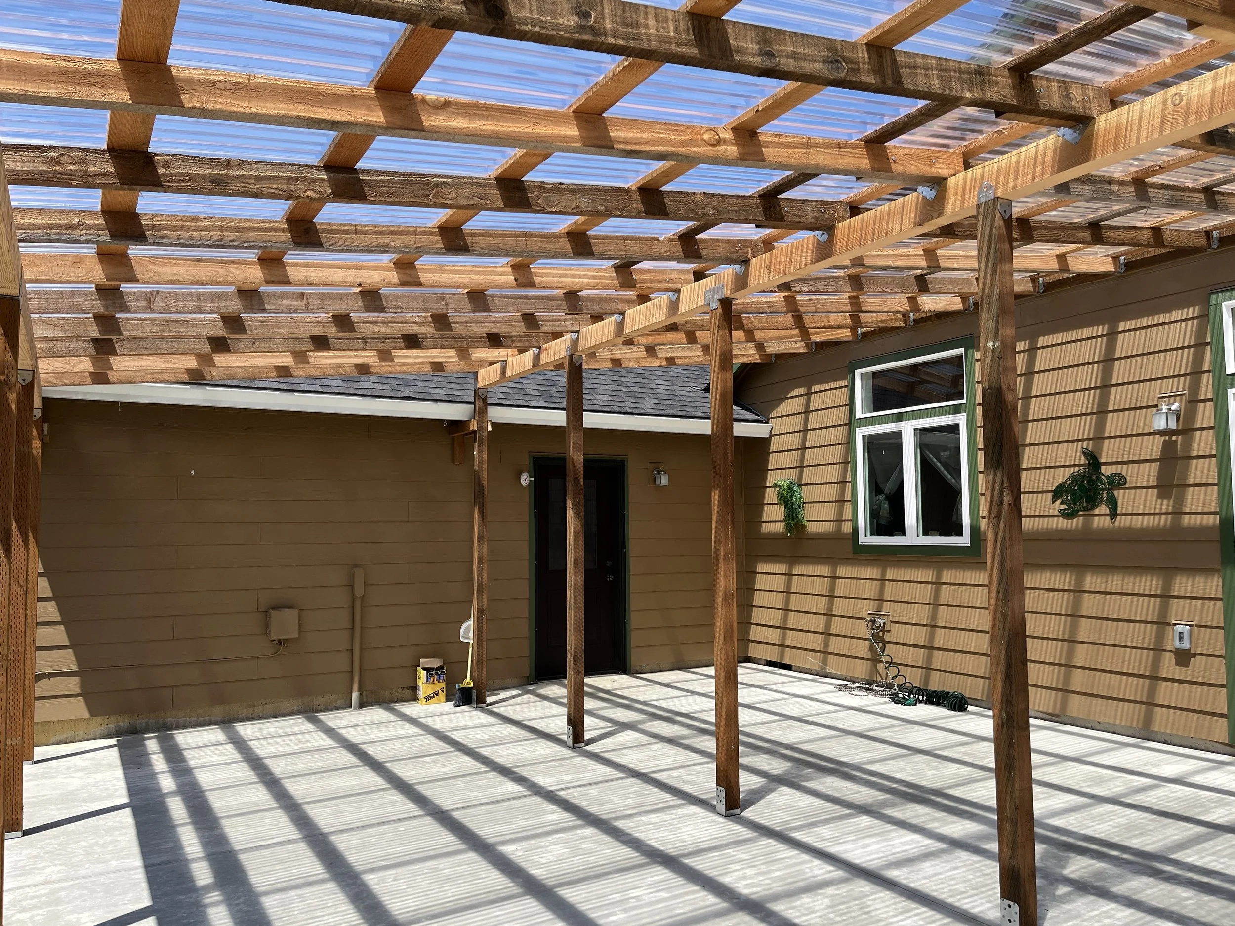 An outdoor construction site with a wooden framework for a roof, supported by vertical wooden posts. The area is enclosed to the side of a house with tan siding, a window, and a door. The ground is concrete, and tools are visible on the floor.