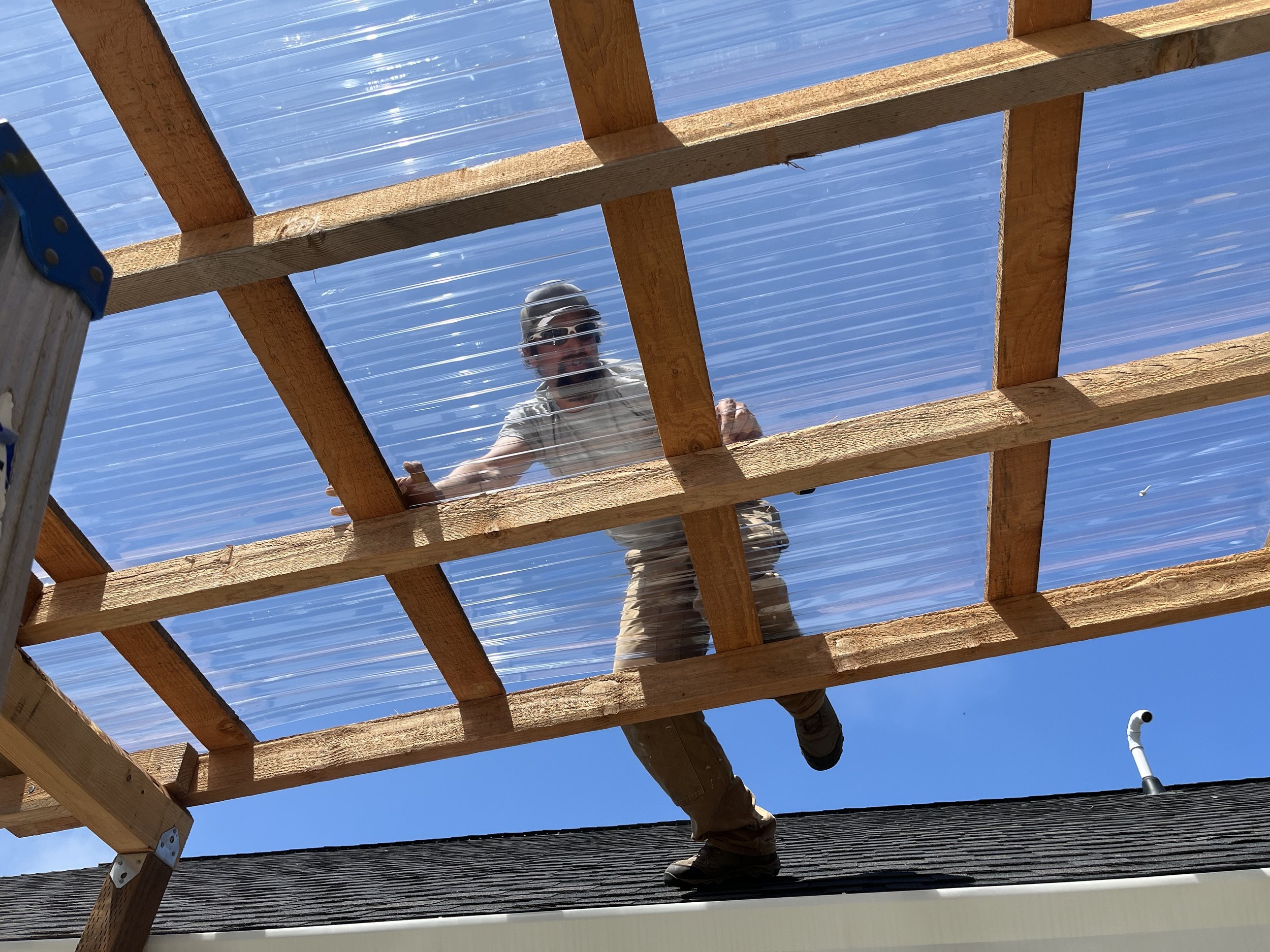 Person working on a wooden roof frame during daytime, with visible blue sky and a vent pipe in the background.