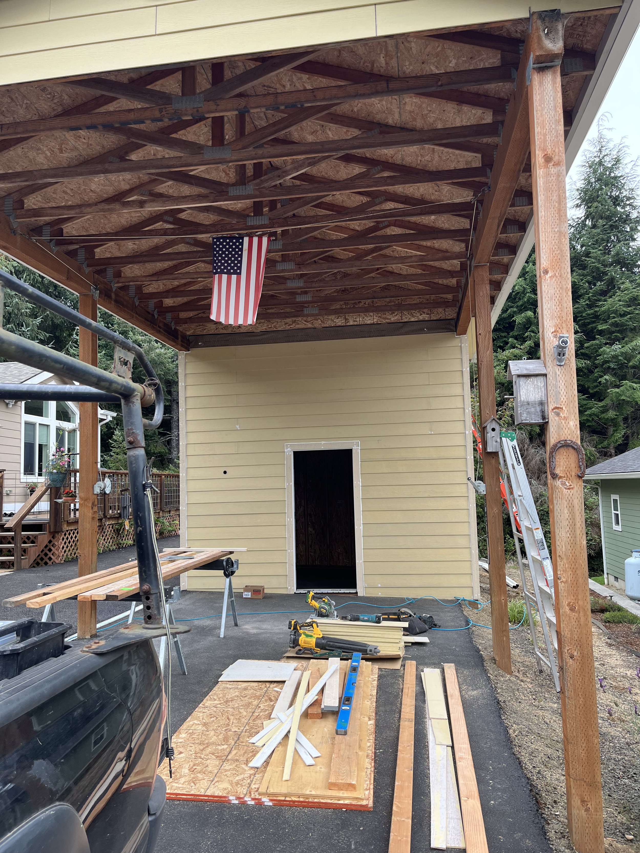 Construction site with a partially built wooden structure. The wall of a small building or shed is visible, and there are various tools and wooden planks on the ground. An American flag is hanging under the roof framing, which consists of wooden beams and trusses. A ladder and some construction materials are also present.
