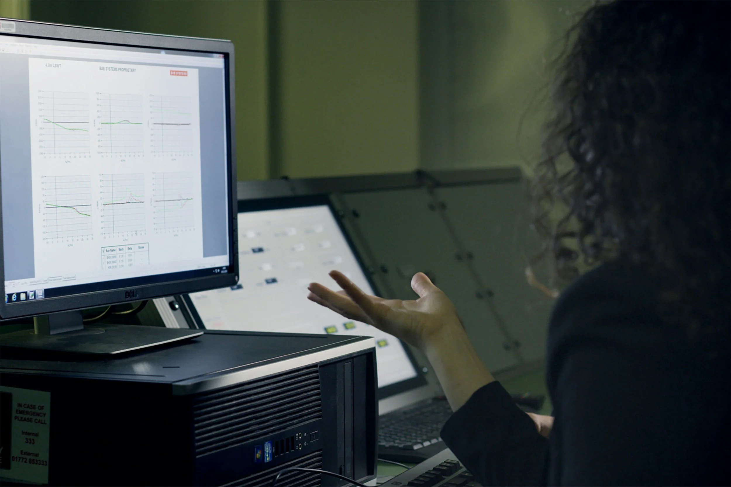 Person with curly hair working on a computer in a control room, monitoring graphs and data on the screen.
