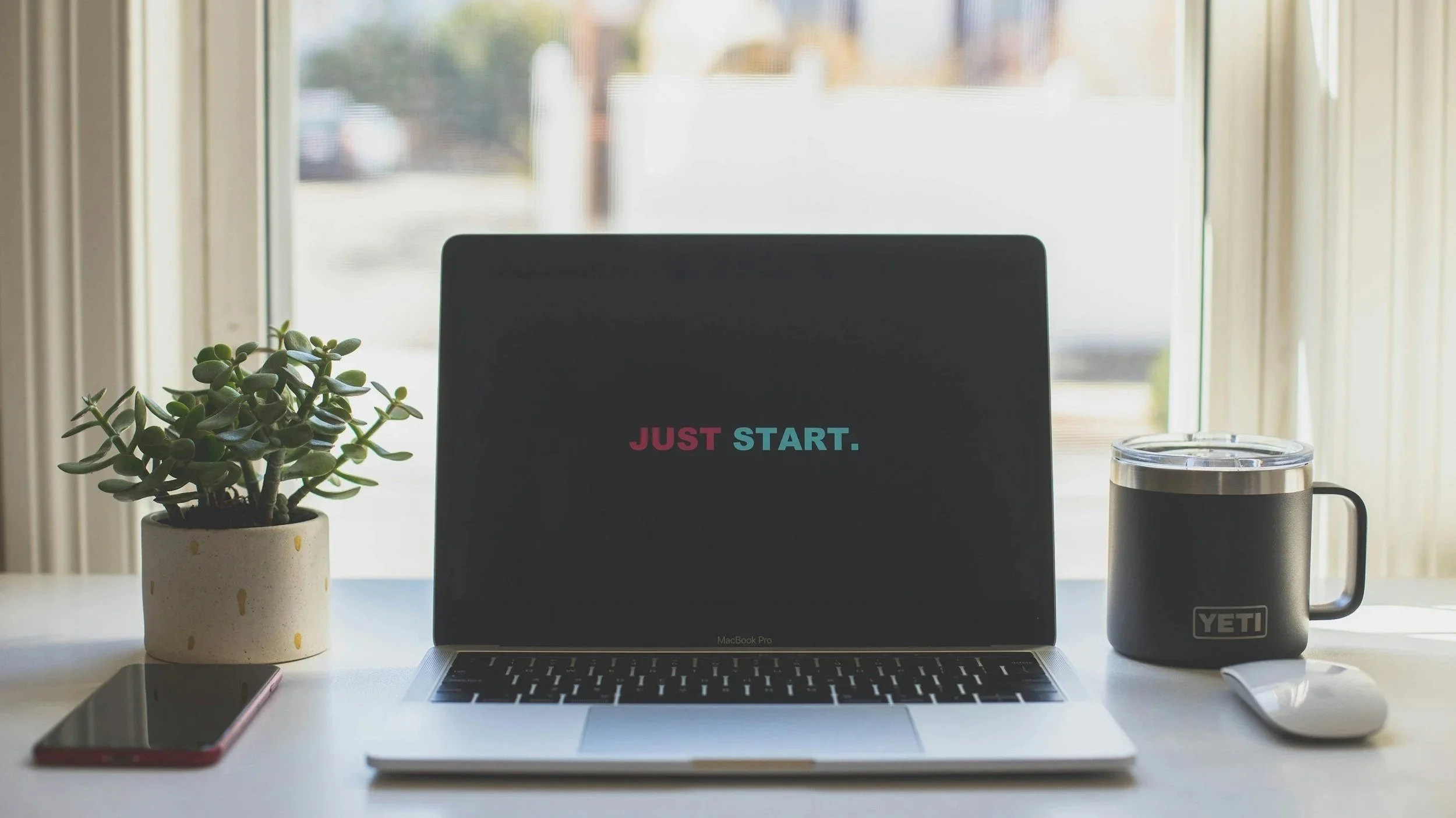 A workspace with a MacBook Pro displaying the words 'JUST START.' in colorful text, a potted succulent, a smartphone, a YETI mug, and a clear tumbler with a black lid on a white table near a window.
