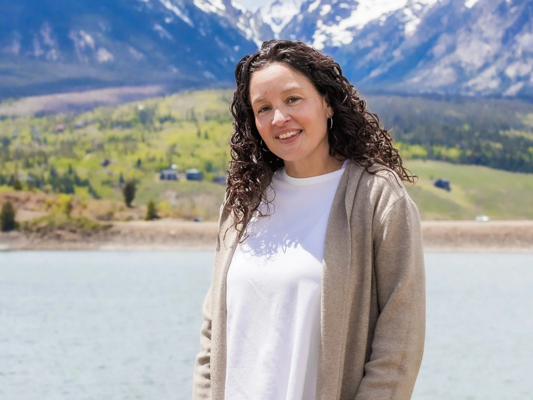 A woman with curly hair smiling outdoors near a body of water with mountains and greenery in the background.