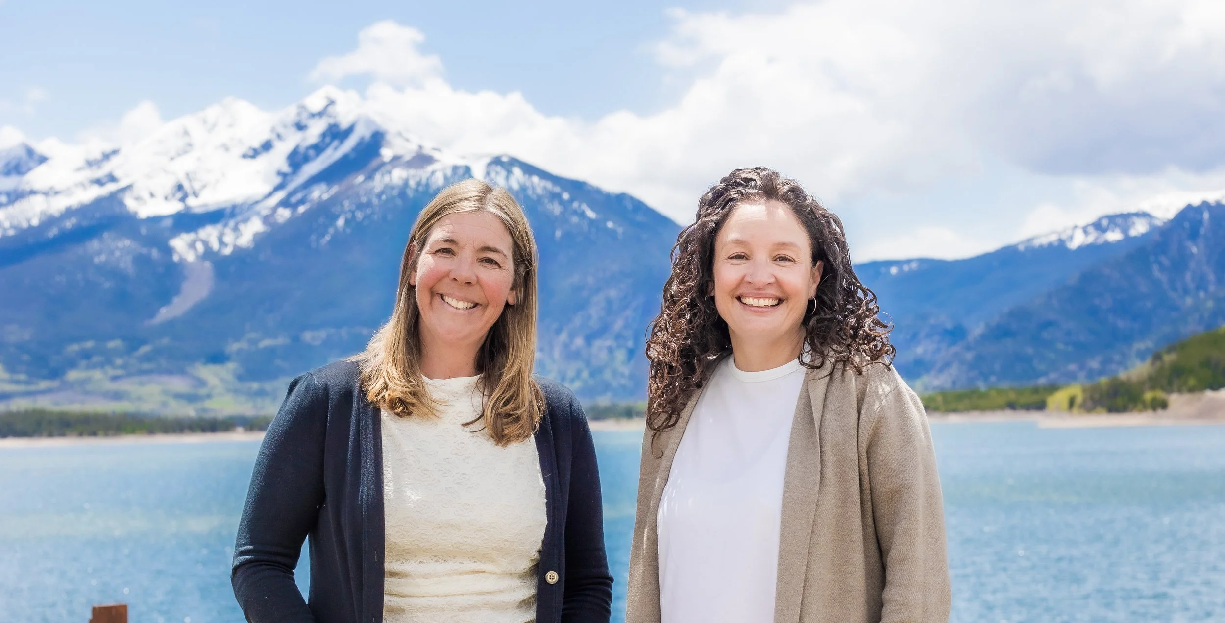 Two smiling women standing outdoors by a lake with snow-capped mountains in the background.