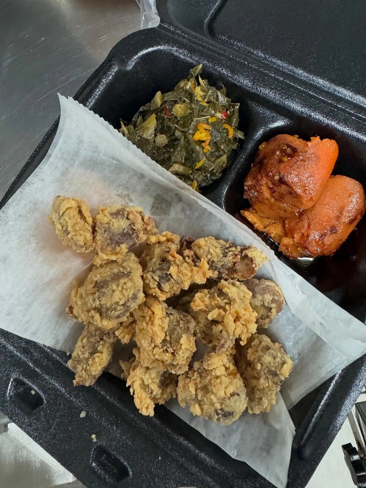 Fried chicken pieces with a crispy golden-brown coating on a black tray, with paper lining, and compartments containing cooked greens and two cooked, seasoned chicken drumsticks.