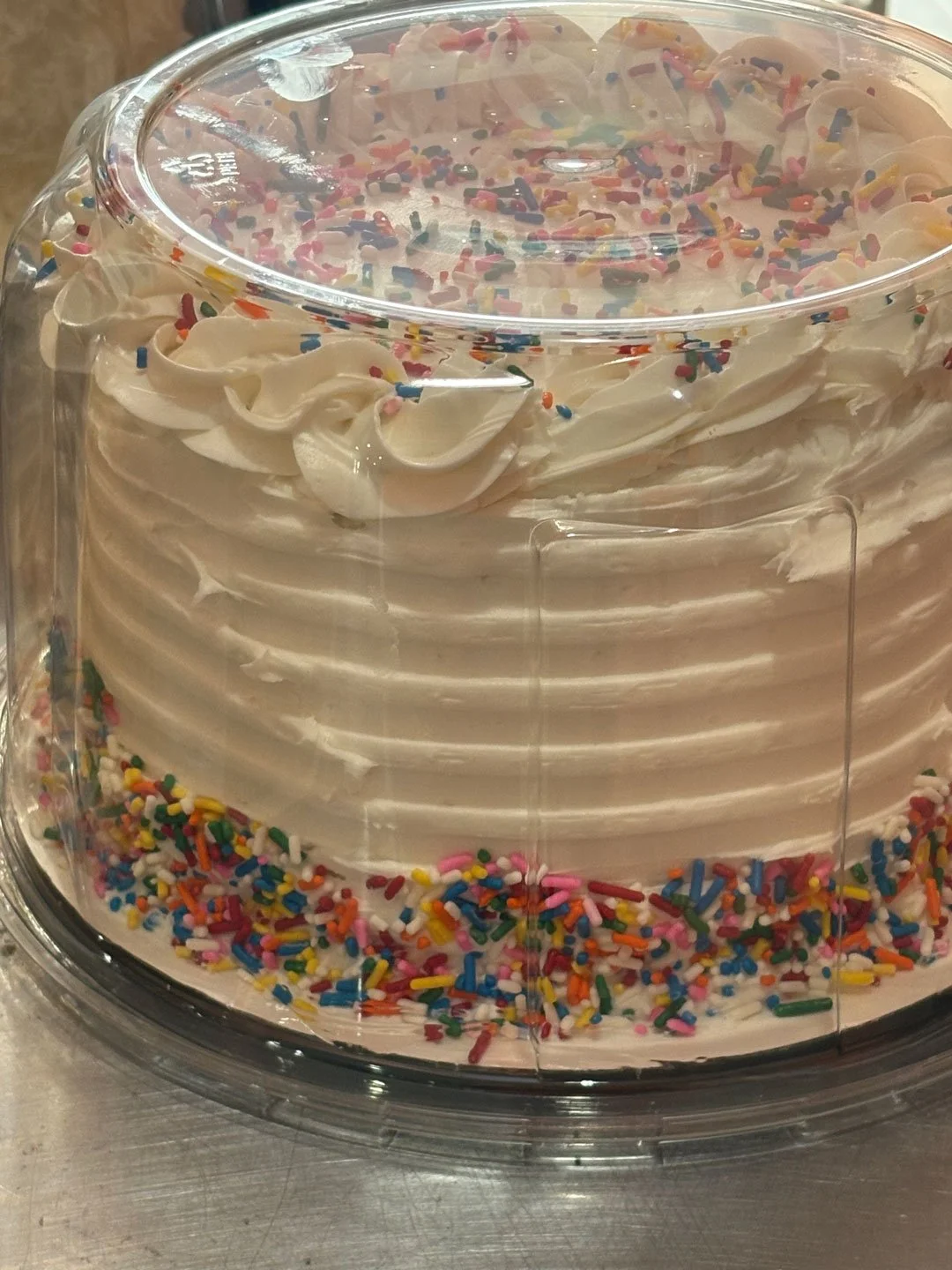 A round cake with white frosting and colorful sprinkles on the top and bottom edges, covered with a clear plastic dome.