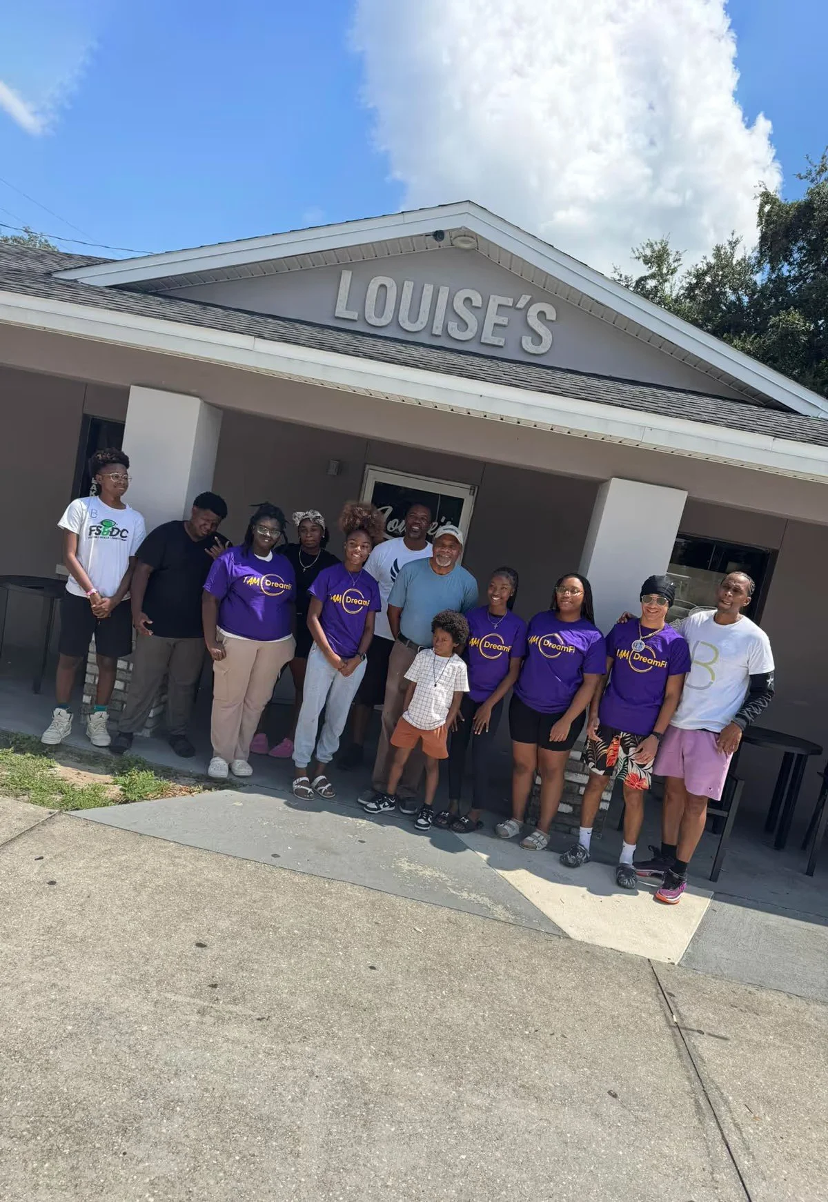 Group of people standing in front of a building with a sign that reads 'Louise's.' Some are wearing purple shirts with logos, and others are in casual attire. The sky is partly cloudy.