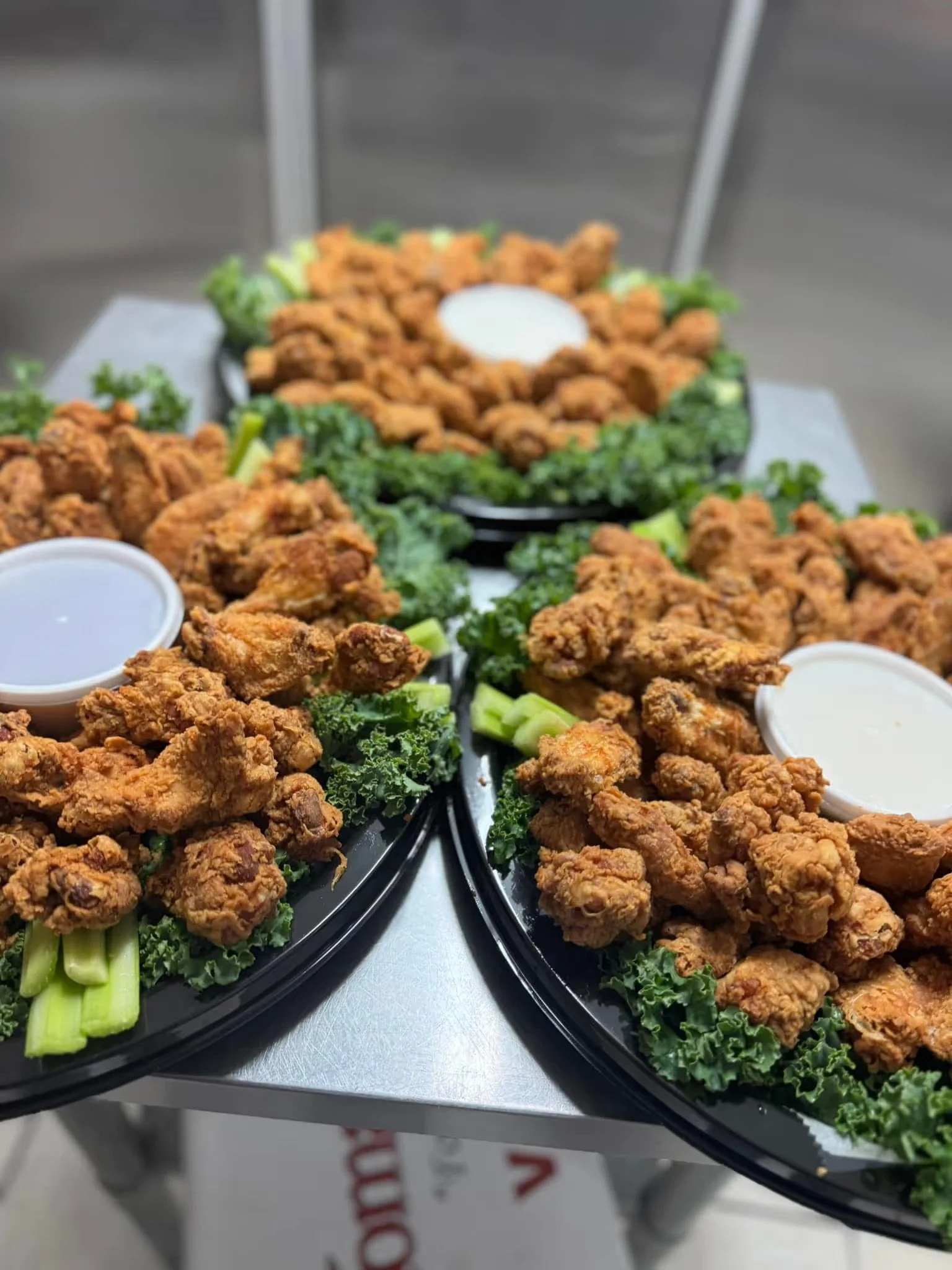 Several black trays filled with fried chicken pieces garnished with green celery sticks and curly kale, with small containers of dipping sauces, on a stainless steel counter.