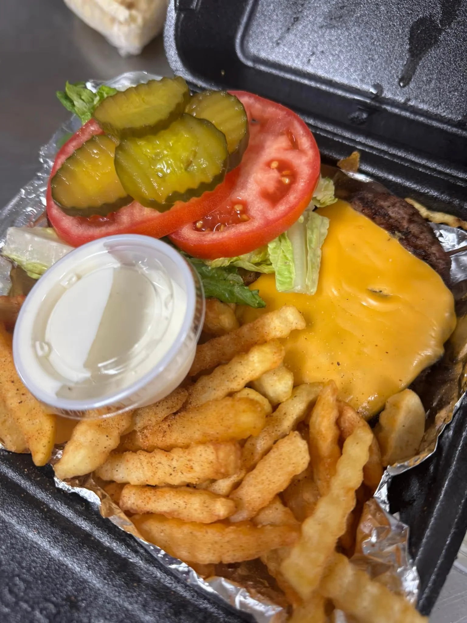 A loaded cheeseburger with lettuce, tomato, pickles, melted cheese, and a side of French fries with a small container of ranch dressing. Grilled meat patty, sizzling from the griddle, partially visible.