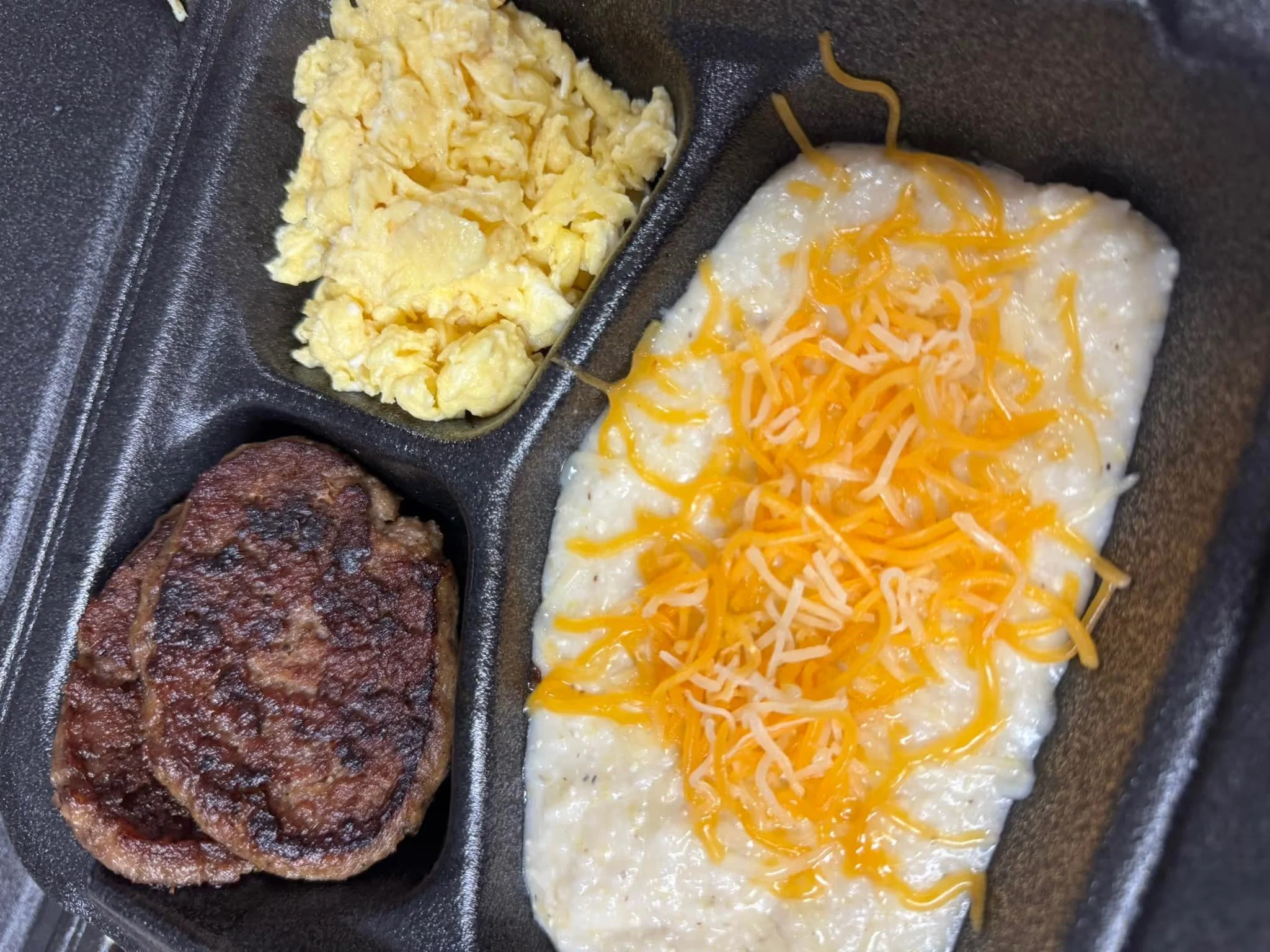 Fried egg with shredded cheddar cheese, cooked scrambled eggs, grilled beef patty, and sausage patty in a black compartmentalized tray.
