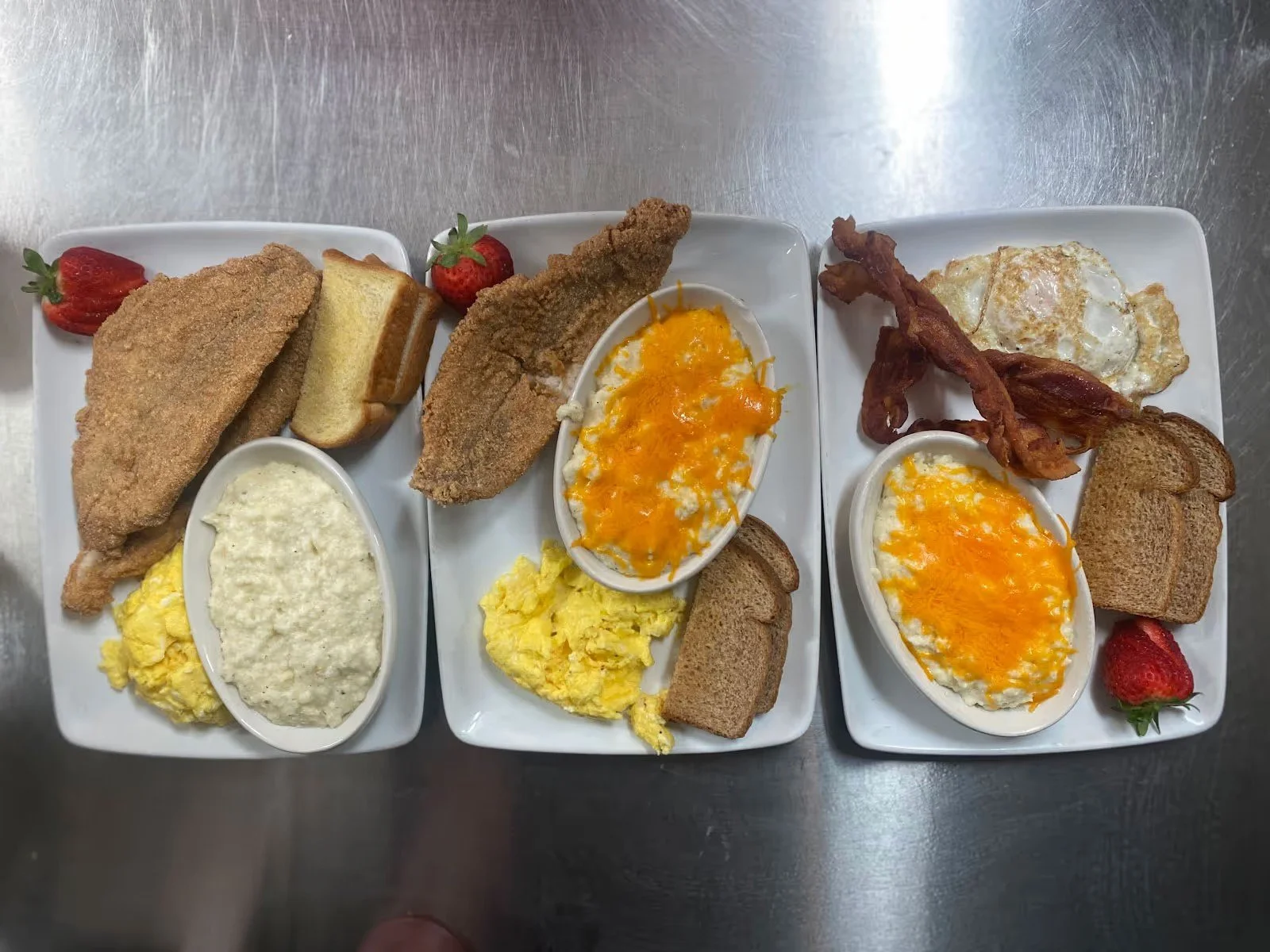 Three breakfast plates with fried fish, scrambled eggs, toast, bacon, and strawberry on a metal countertop.