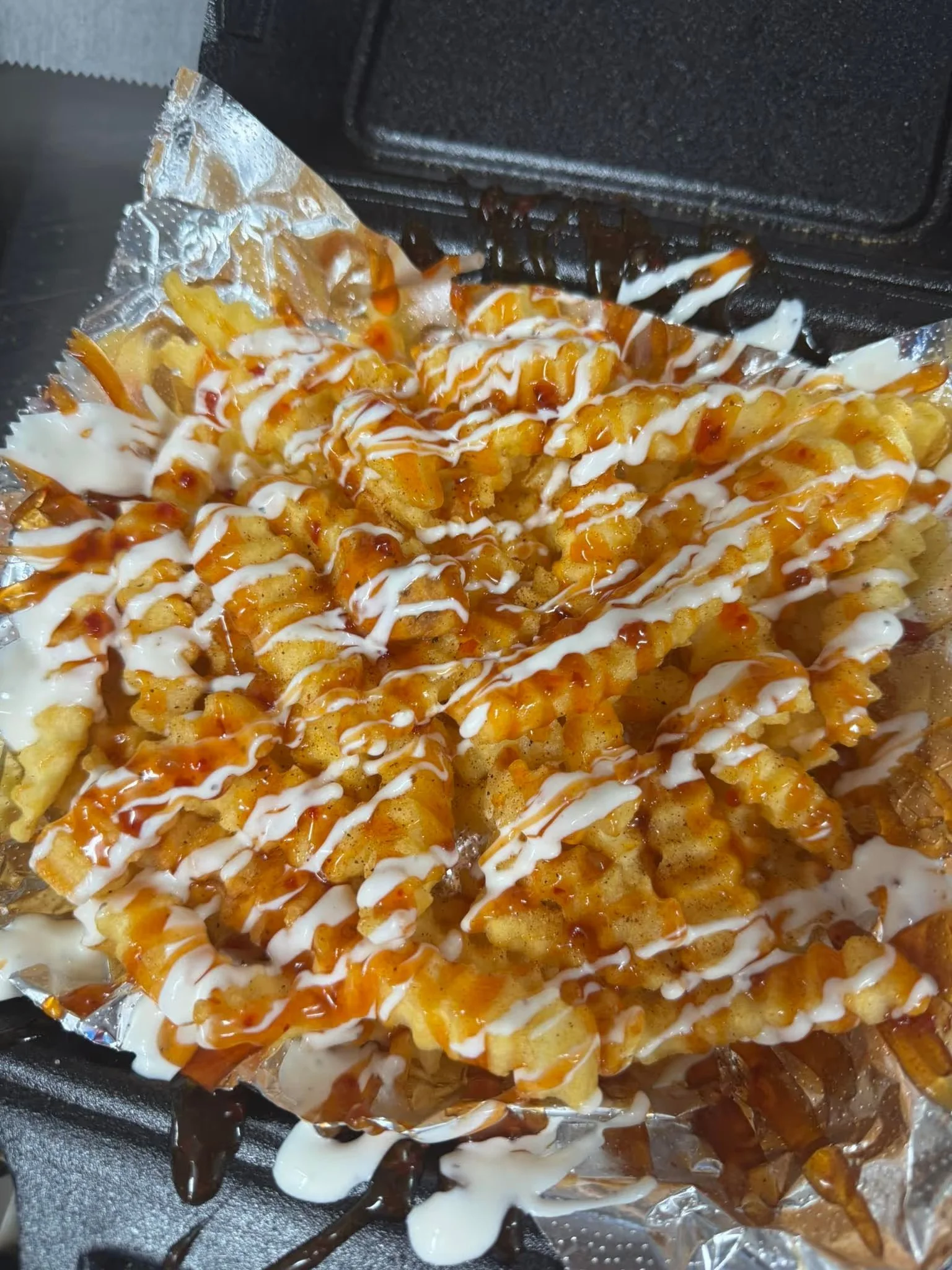 Close-up of crispy French fries topped with drizzles of white mayonnaise and orange ketchup sauce, served in a black tray lined with aluminum foil.
