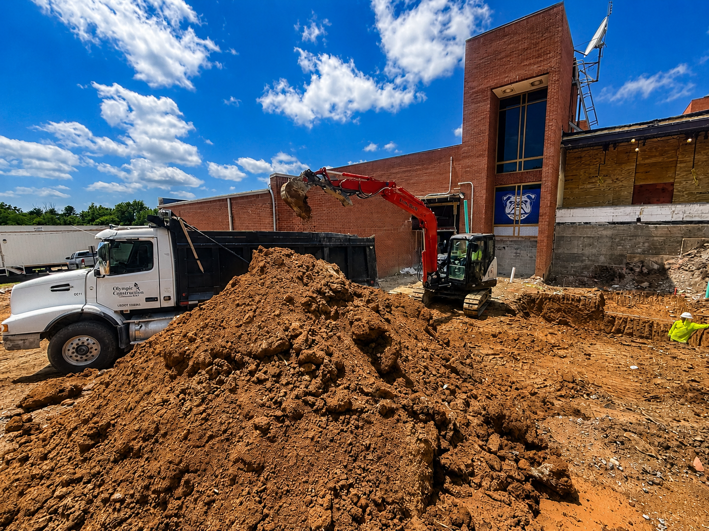 Construction workers and machinery excavate dirt outside a brick building under a partly cloudy blue sky.