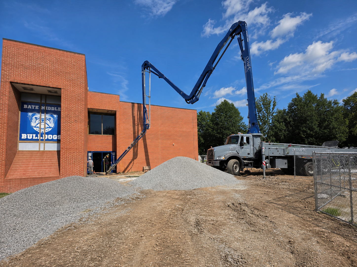 Construction site outside Bate Middle School Bulldogs entrance with a large cement pump truck pouring concrete, piles of gravel, and workers working near the building under a partly cloudy sky.