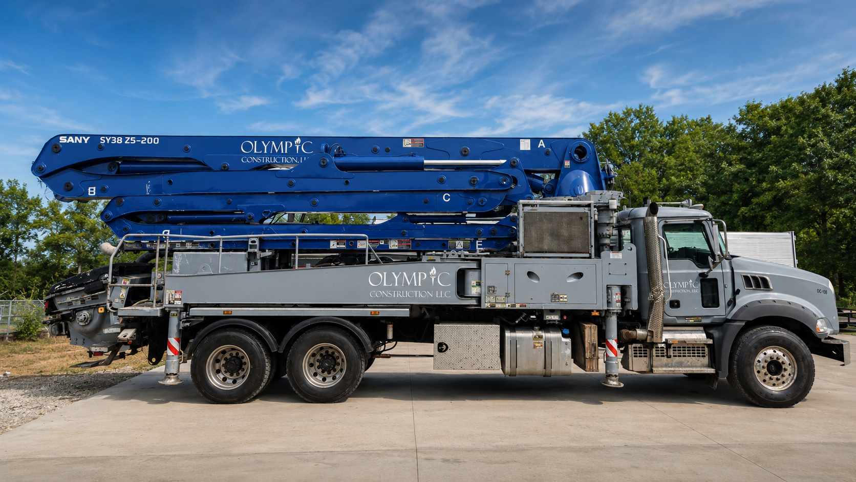 A large construction crane truck with a blue boom and the logo 'Olympic Construction LLC,' parked outdoors on a concrete surface under a blue sky with some clouds, surrounded by green trees.