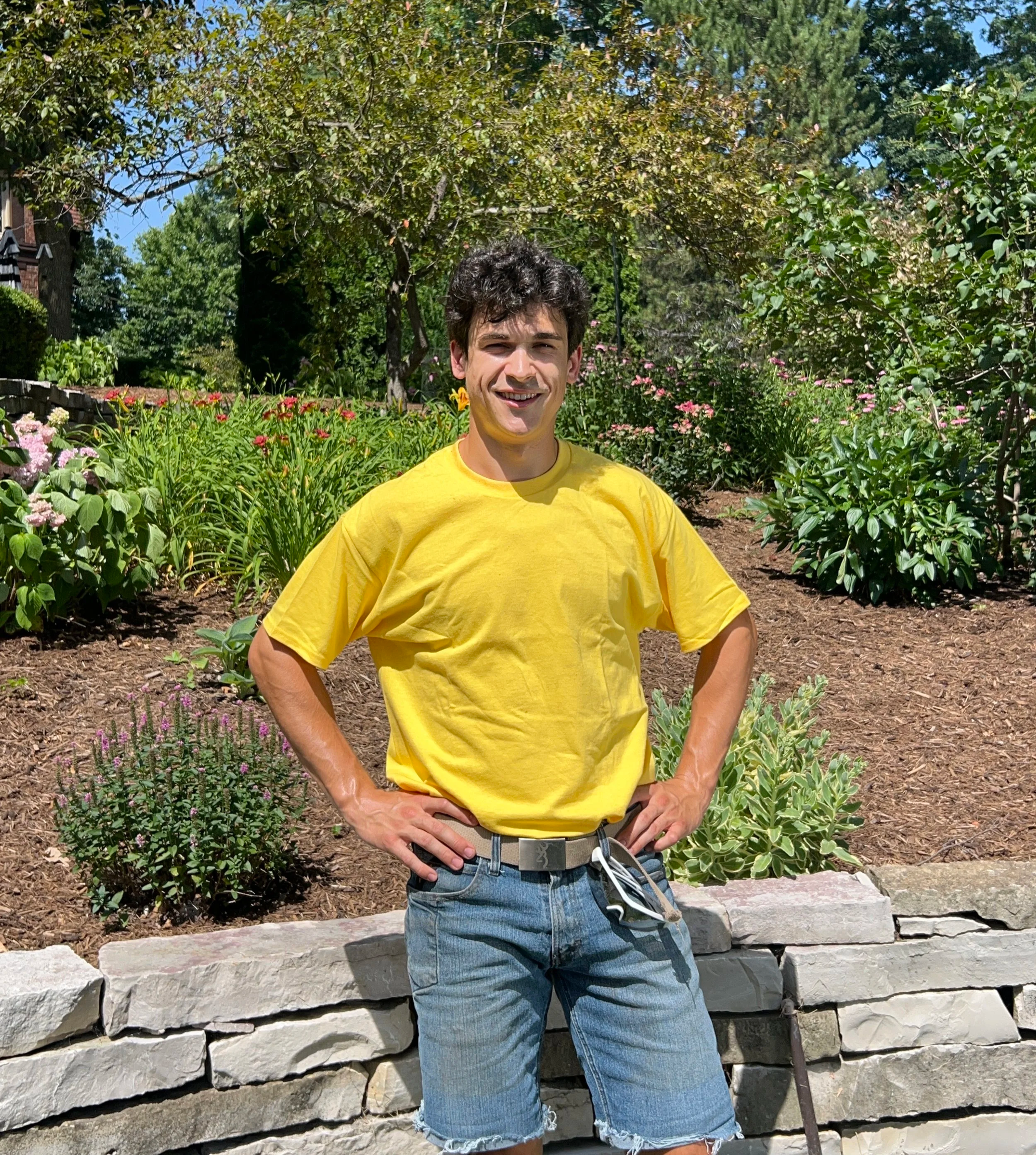 A young man with dark curly hair, wearing a yellow t-shirt and denim shorts, stands outdoors with one hand on his hip, smiling at the camera. He is surrounded by garden flowers, trees, and a stone wall.