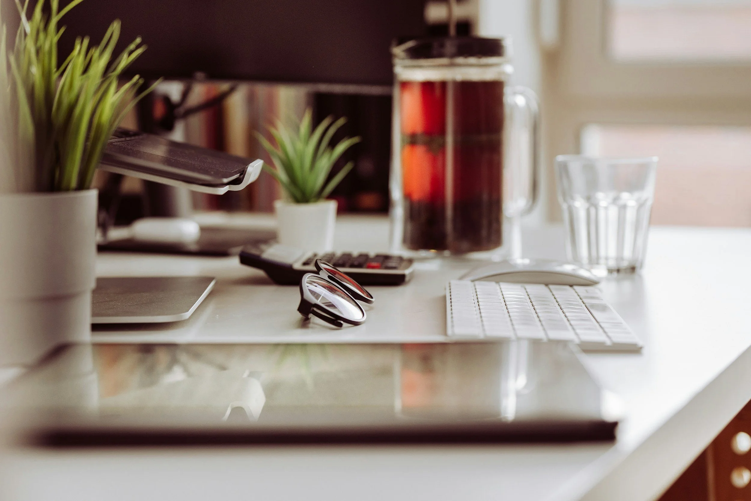 A cluttered white desk with a computer monitor, a small plant in a white pot, a pair of black glasses, a remote control, a notebook, a glass of water, a glass pitcher with a red beverage, and other office supplies, with a window in the background.
