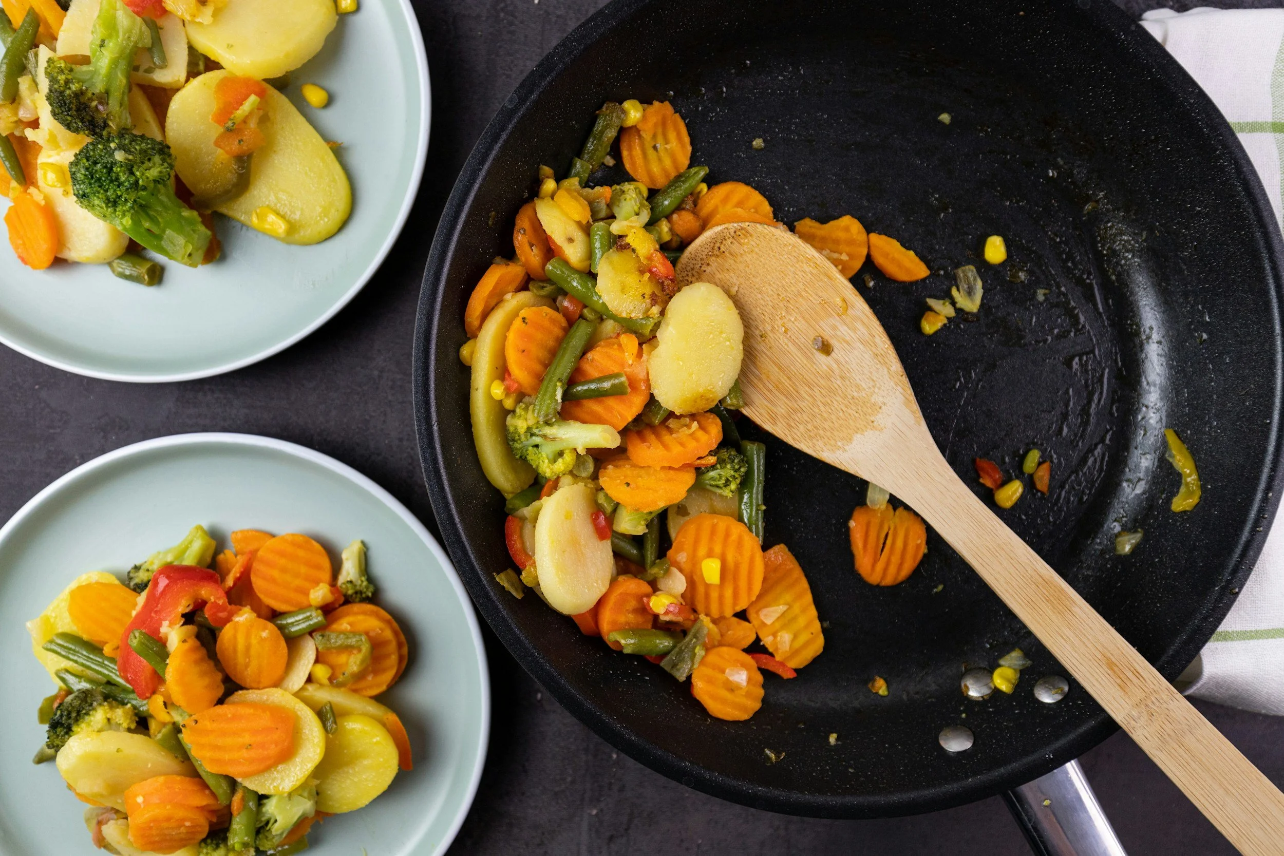 Cooked mixed vegetables including carrots, green beans, broccoli, and potatoes in a frying pan with a wooden spatula, next to plates of cooked vegetables.