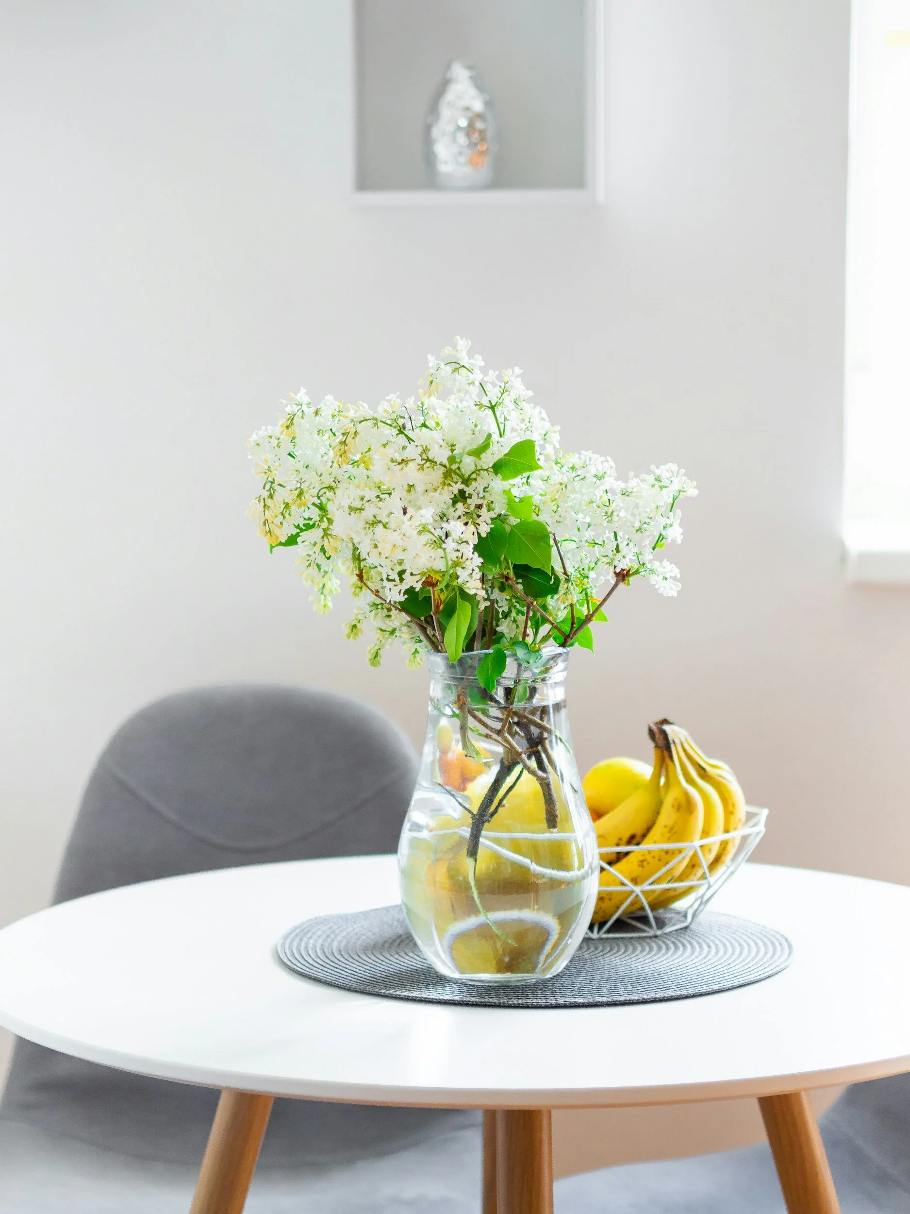 A round white table with wooden legs holds a clear glass vase with white flowers and leaves, and a wire basket filled with bright yellow bananas on a gray circular placemat in a minimalistic setting with white walls and a gray upholstered chair.
