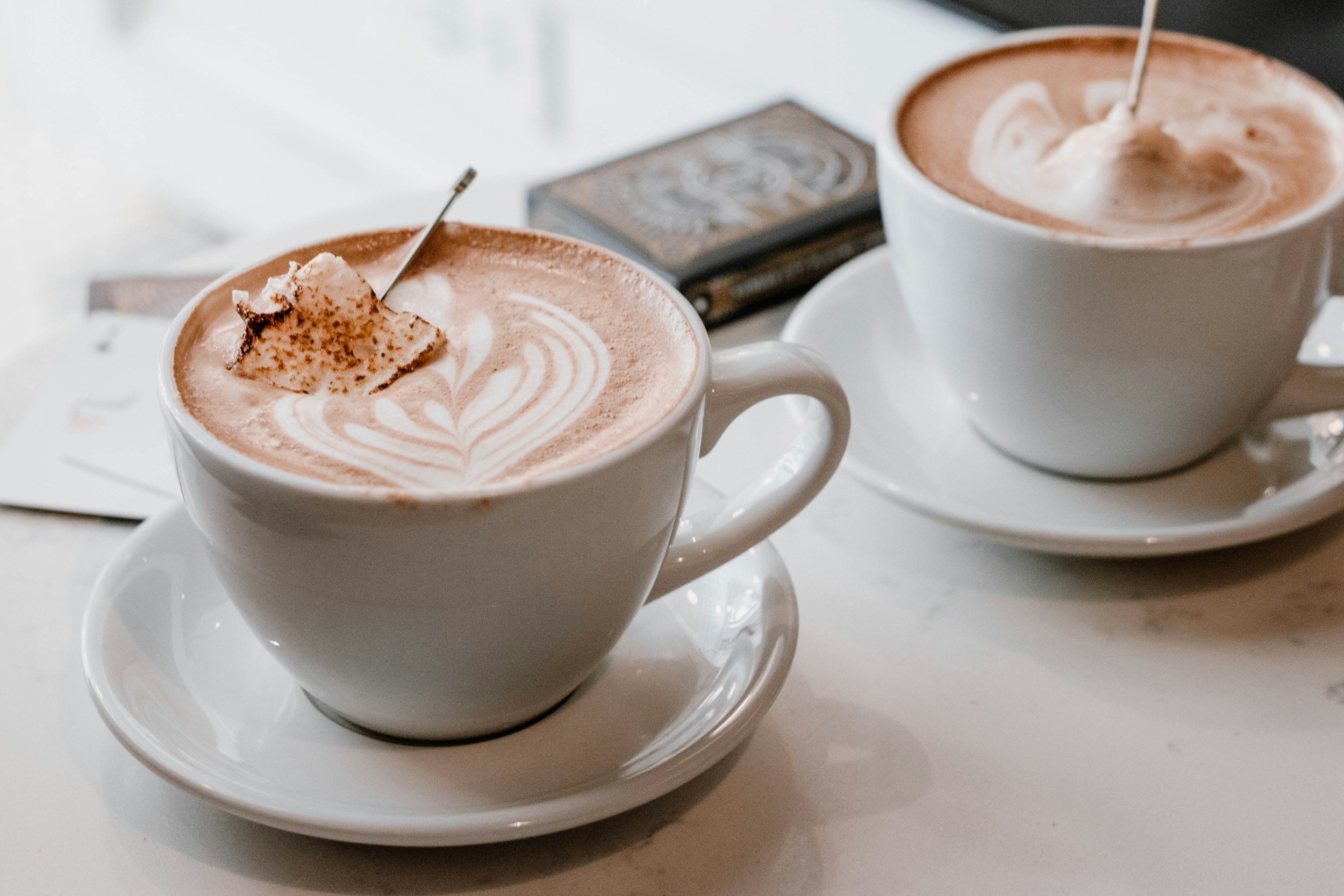Two white coffee cups filled with hot chocolate, topped with whipped cream and a sprinkle of cocoa powder, sit on matching saucers on a white surface.