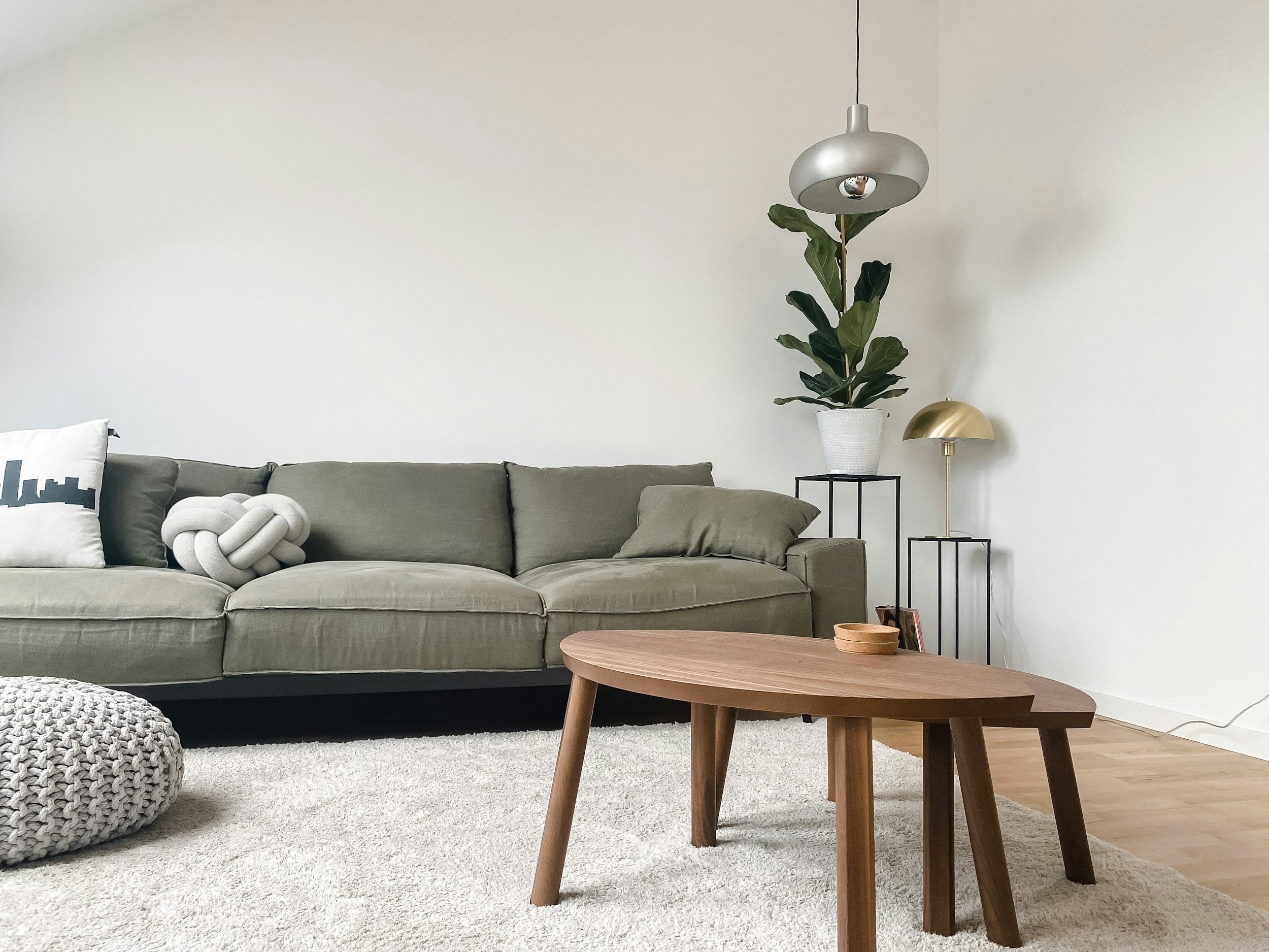Living room with a gray sofa, decorative pillows, a chunky knitted pouf, a wooden coffee table, a potted plant, a gold table lamp, and a hanging silver lamp against a plain white wall.
