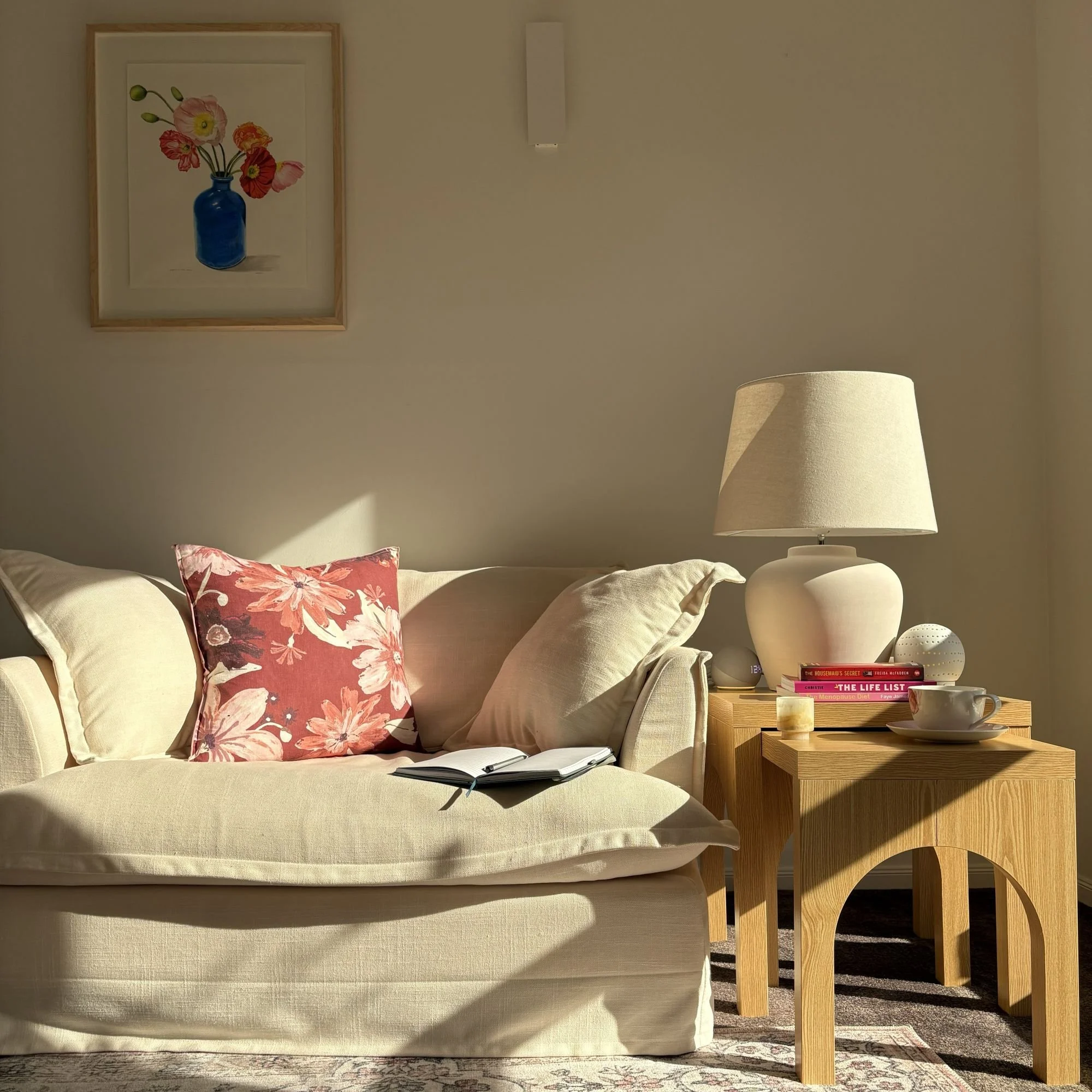 A cozy bedroom corner with a beige upholstered bed, floral pillow, wooden nightstand with books and a lamp, wall art of a blue vase with flowers, and a cream-colored lampshade.