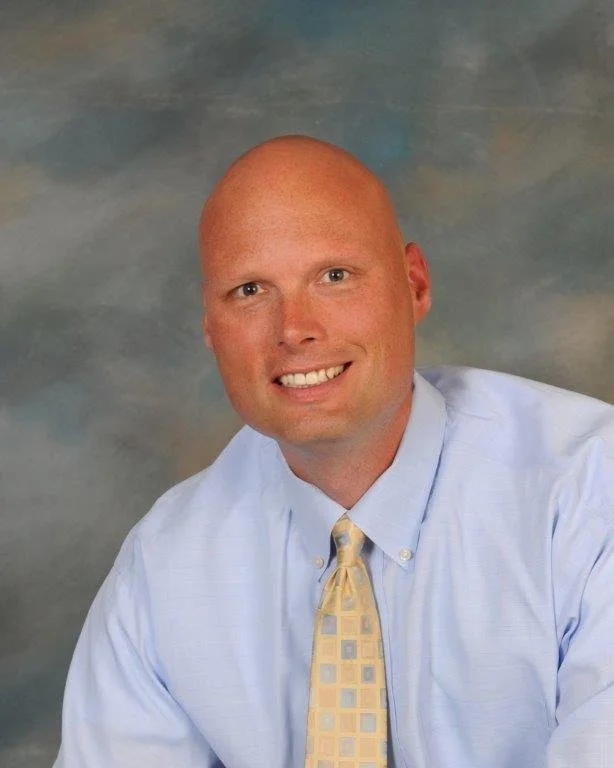 A man with a bald head wearing a light blue dress shirt and a yellow checkered tie, smiling against a cloudy background.