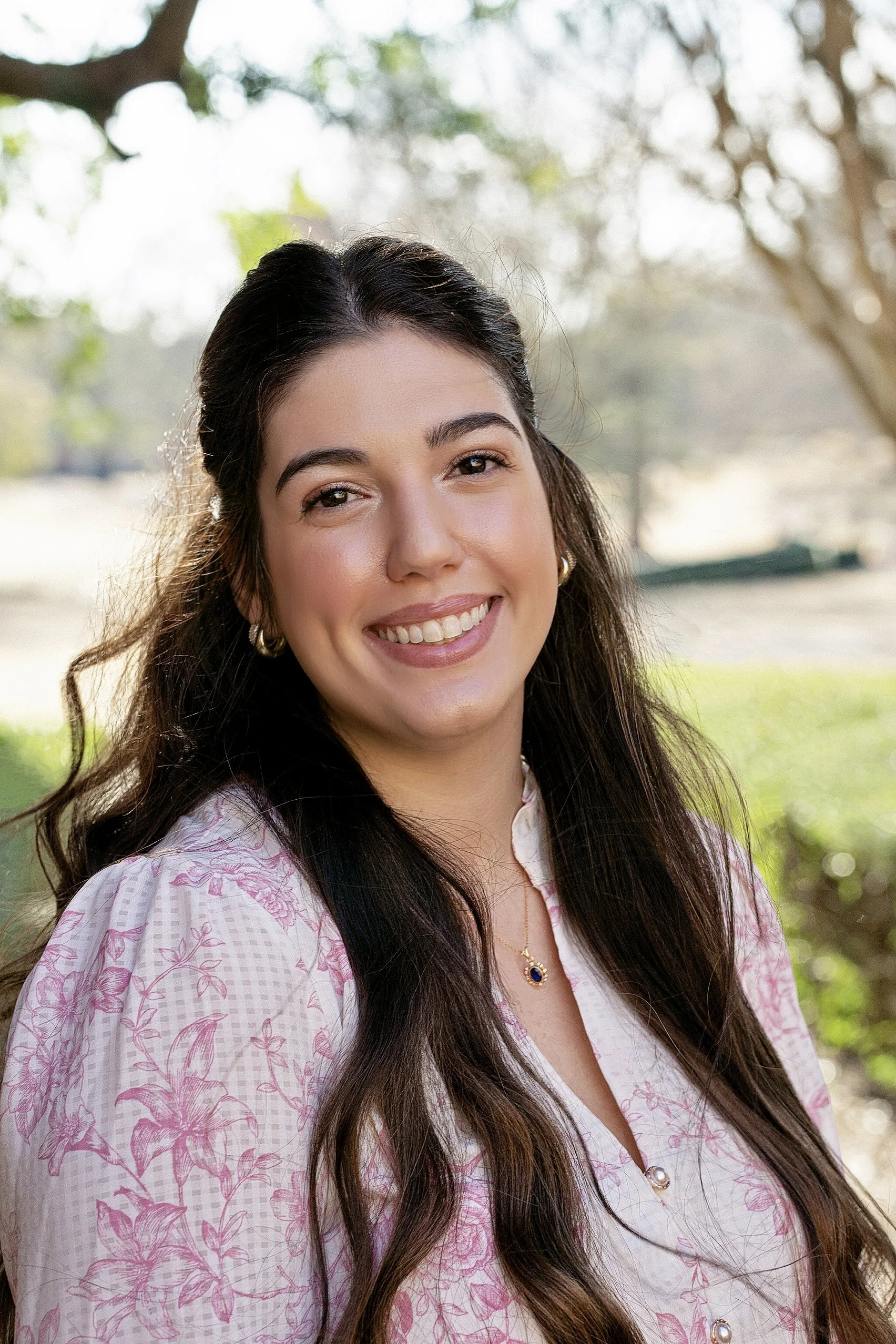 A smiling woman with long dark hair and gold hoop earrings outdoors in a park with trees and greenery in the background.