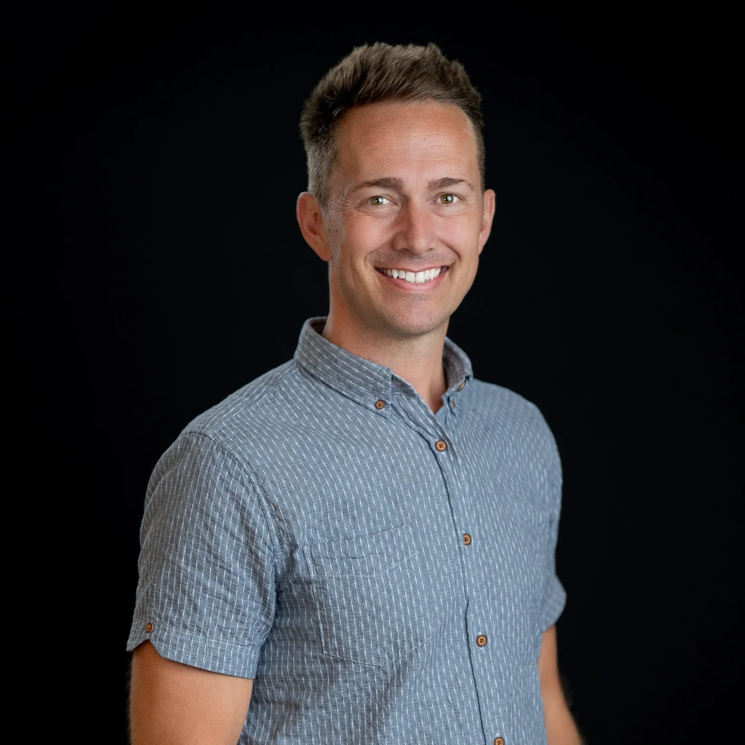 A smiling man with short brown hair and a light beard wearing a blue patterned short-sleeve button-up shirt against a black background.