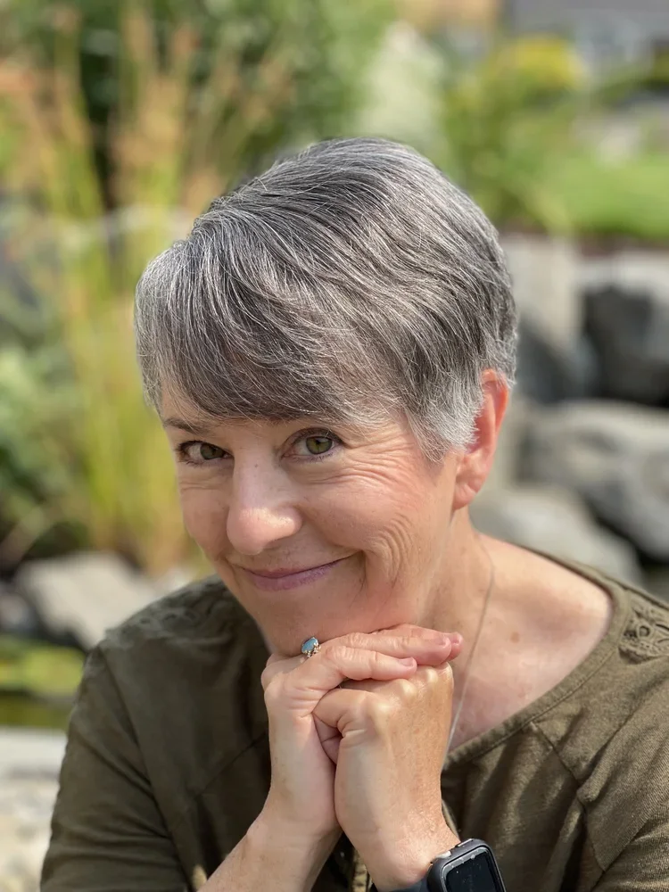 A smiling middle-aged woman with short gray hair, wearing a green top, outdoors with greenery and rocks in the background.