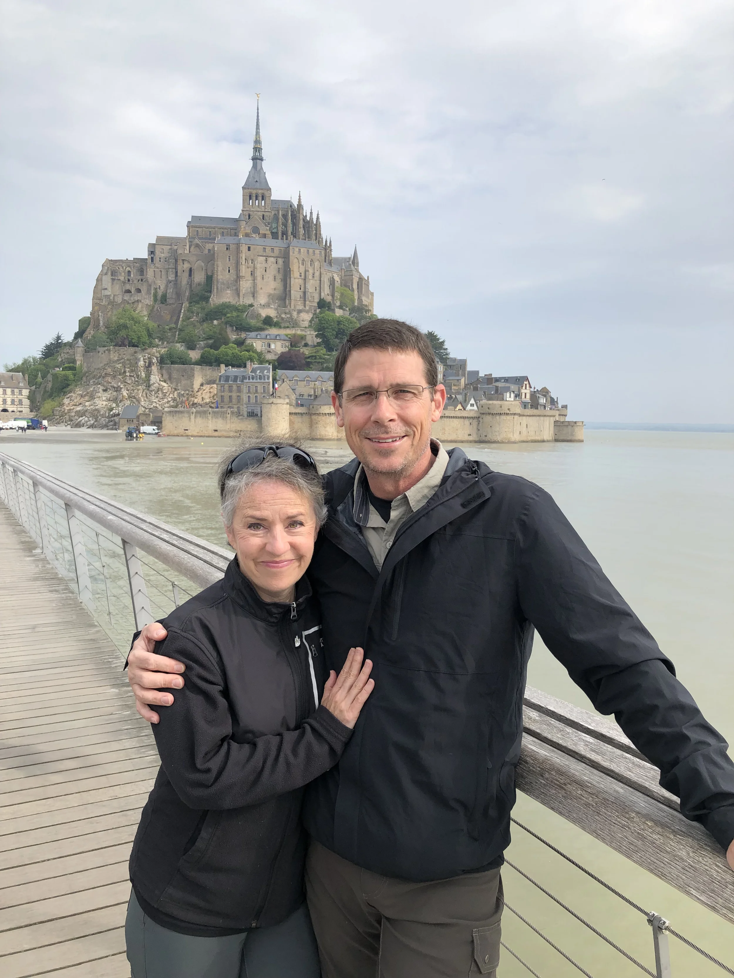 A man and woman hugging on a wooden bridge with a castle on a hill in the background during cloudy weather.