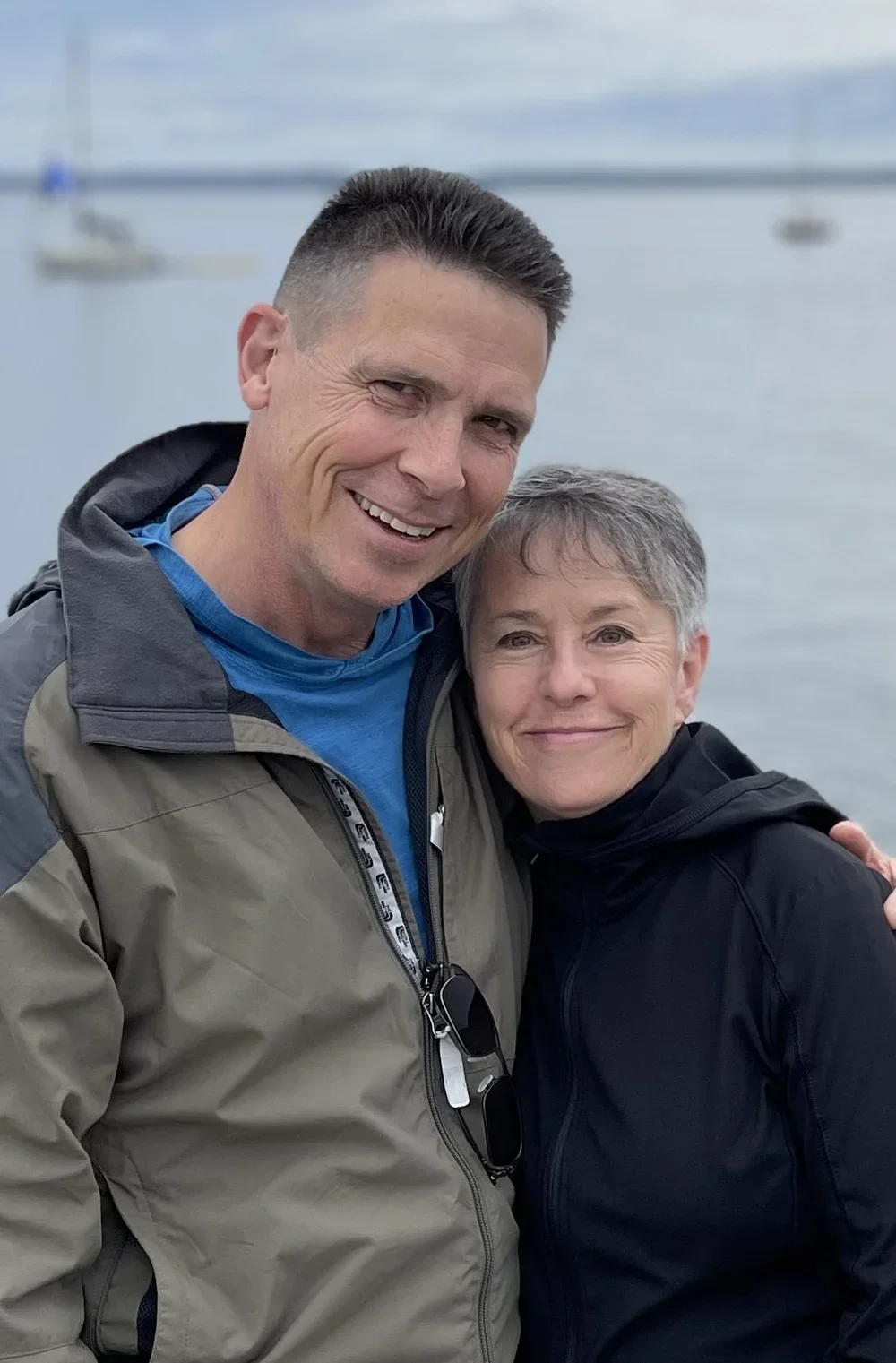 A man and woman smiling and hugging near a body of water with sailboats in the background.