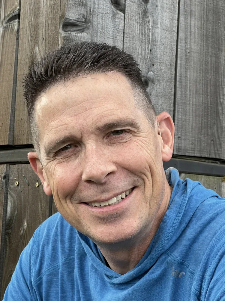 A smiling man with short dark hair, wearing a blue shirt, sitting outdoors in front of a wooden fence.