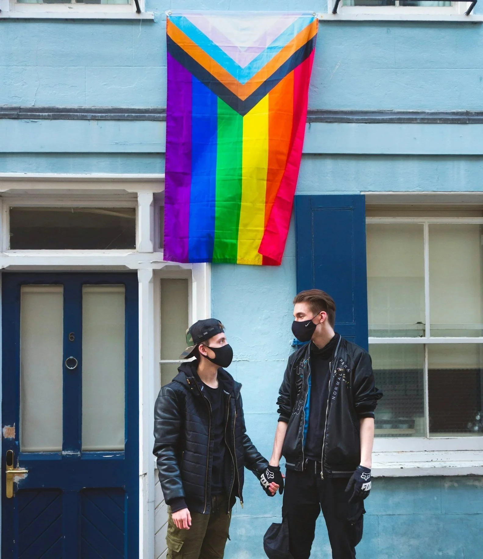 Two young men are standing outside, wearing black face masks and black jackets, holding hands and looking at each other. Behind them is a blue door and a LGBTQIA+ pride flag hanging above them.