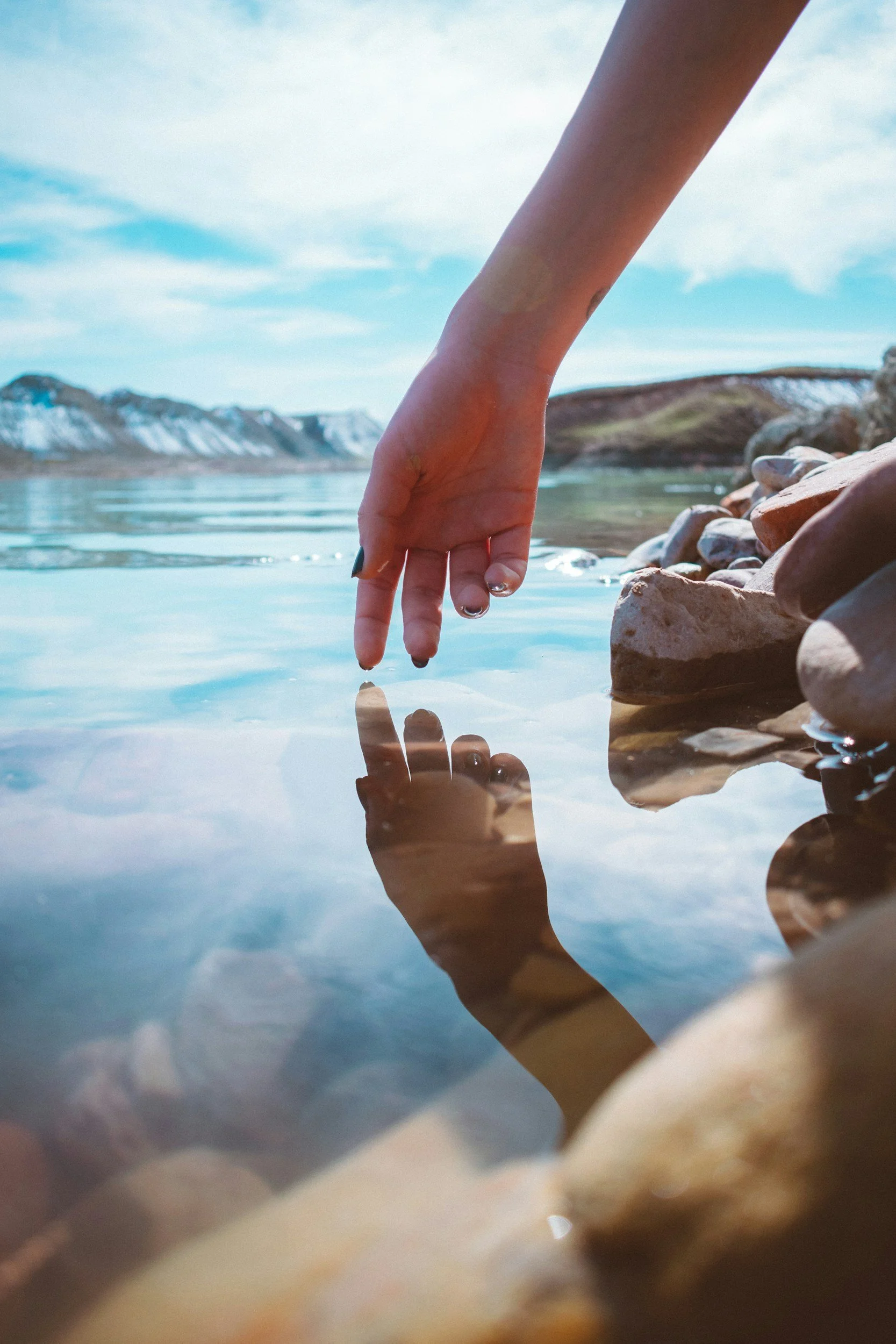 Close-up of a hand and fingers touching the water's surface with a mirror image reflecting in the water on a rocky shoreline, with mountains and a cloudy sky in the background.