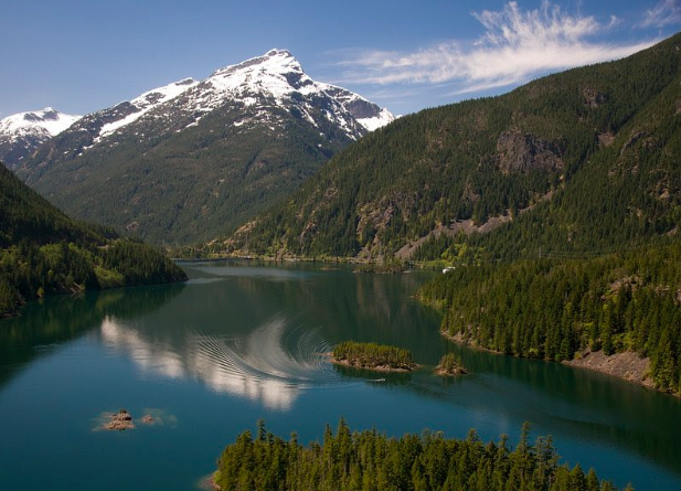 A calm lake surrounded by lush green forests with a snow-capped mountain in the background, under a partly sunny sky.