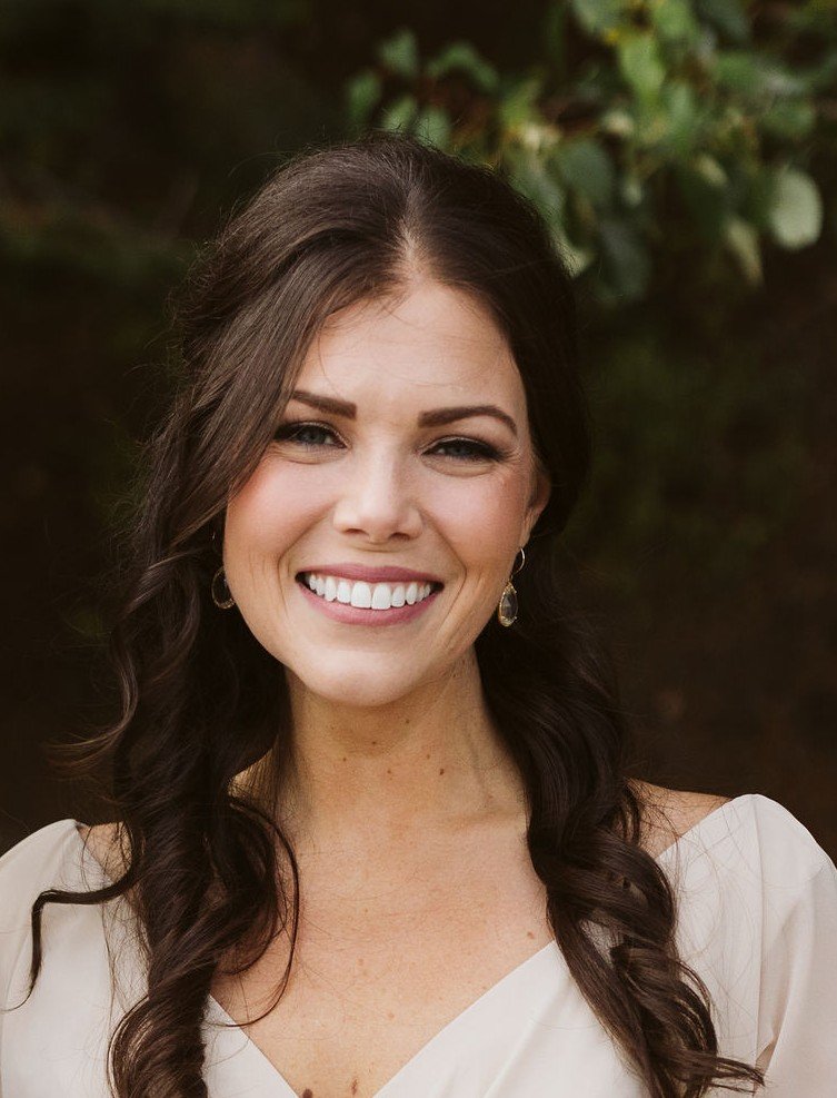 Alexx Ward, therapist, a young woman with long brown hair, smiling, wearing a white top and earrings, outdoors with greenery in the background.