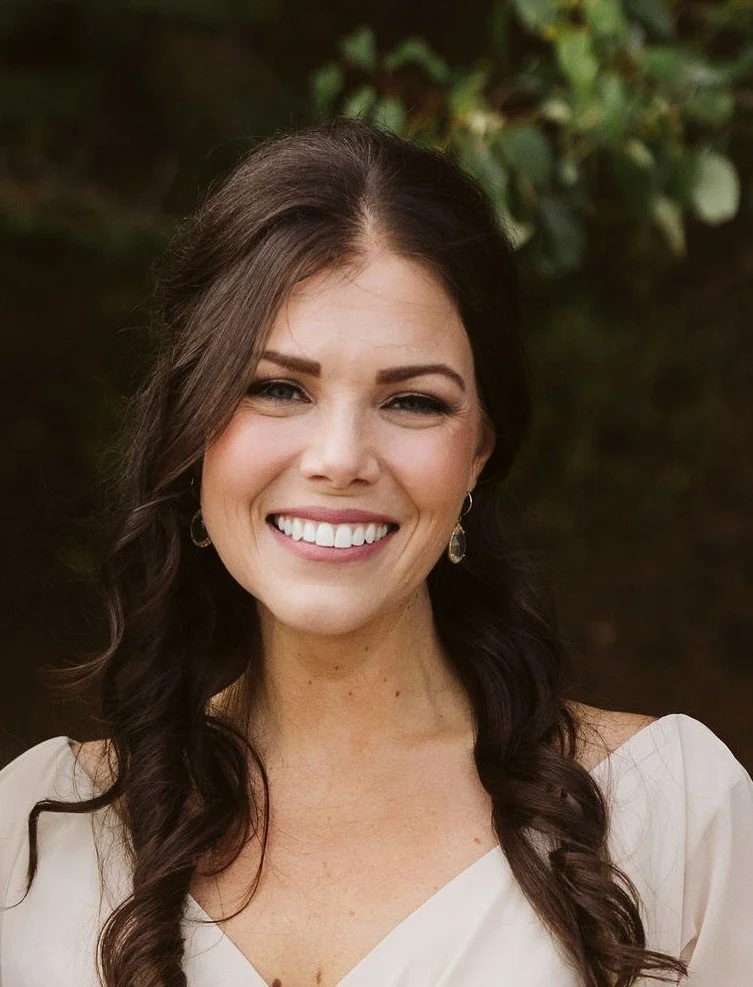 Alexx Ward, therapist, a young woman with long brown hair, smiling, wearing a white top and earrings, outdoors with greenery in the background.