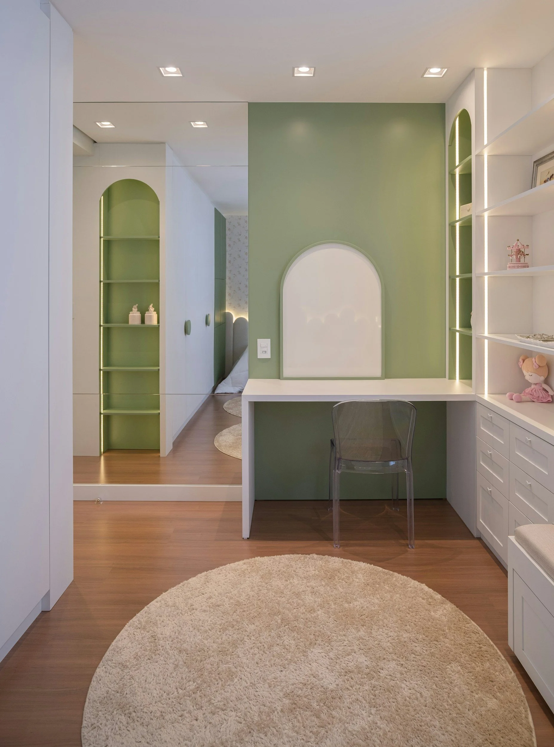 Interior view of a room with wooden flooring, a circular beige shaggy rug, and built-in white cabinetry with shelves. The room features a green accent wall with a rounded arch design and integrated lighting, and includes a transparent acrylic chair at a small white desk.
