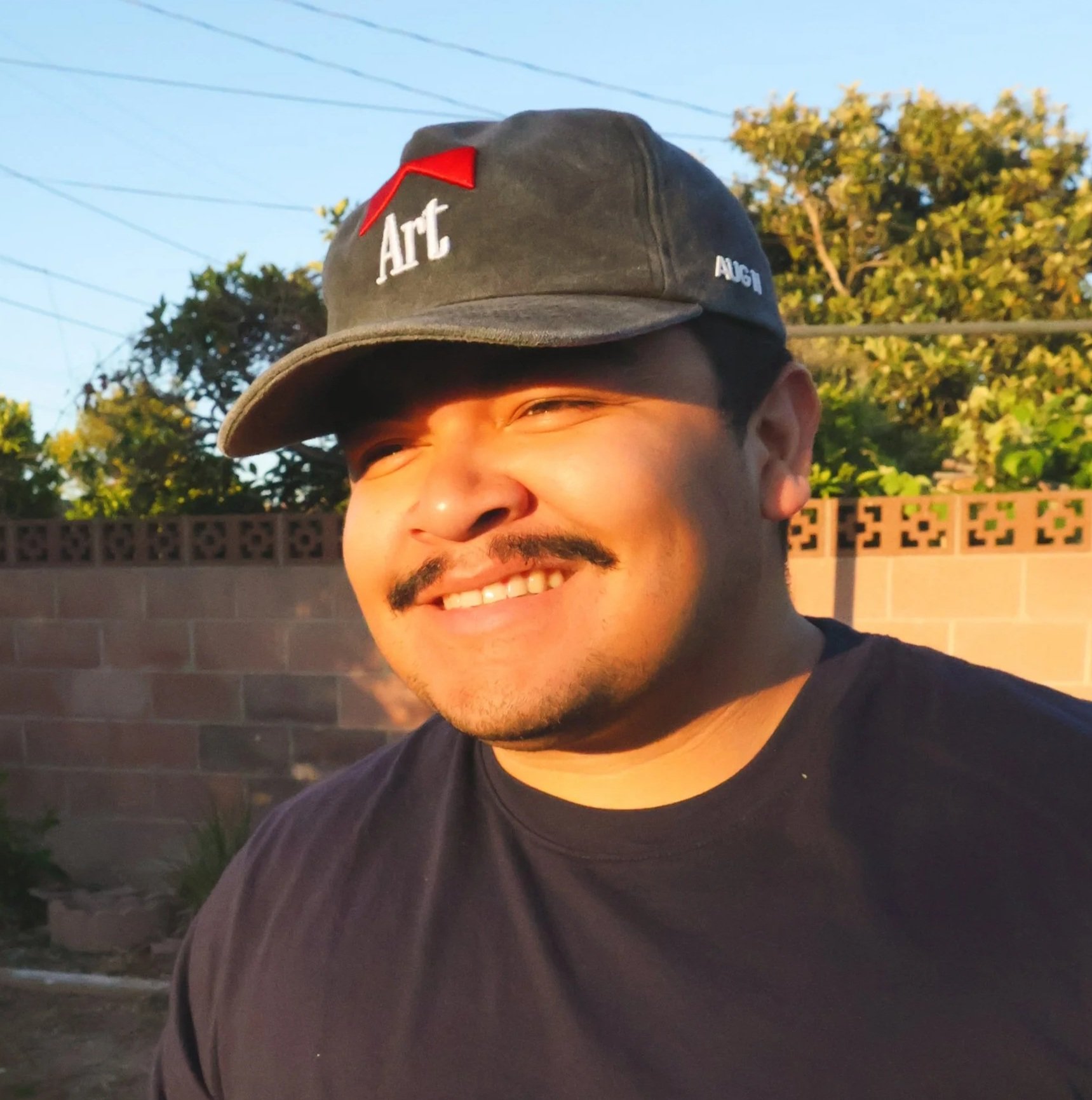 A smiling man wearing a black cap with the word 'Art' embroidered on it and a red arrow, and a black shirt, standing outdoors during sunset with trees, a brick wall, and power lines in the background.