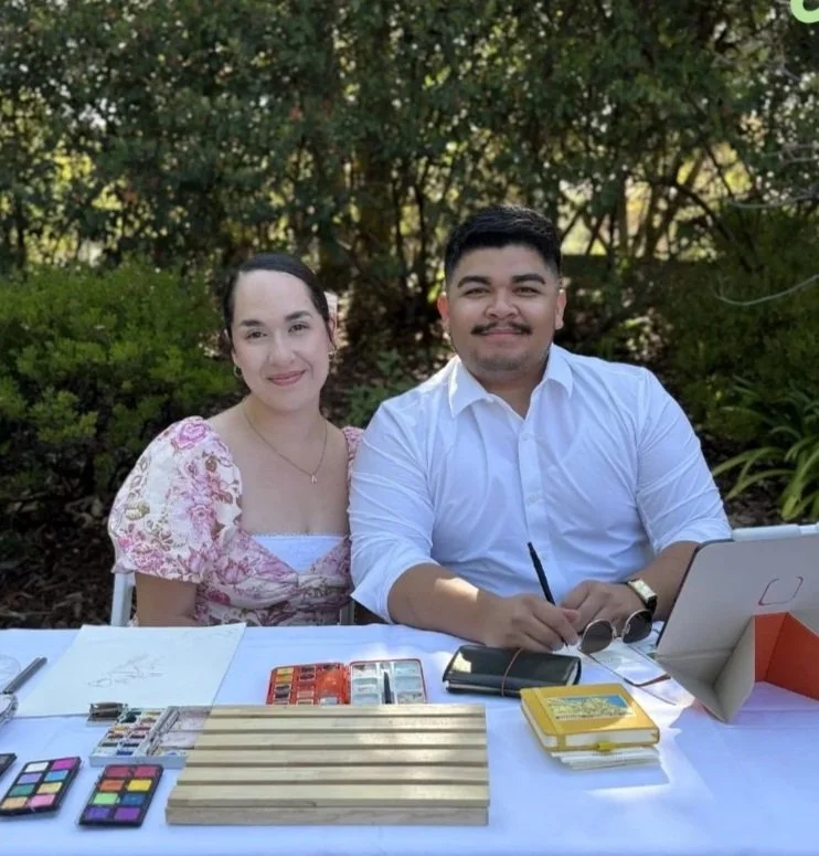 A woman and a man sitting at a table outdoors with makeup palettes, jewelry, and other items, surrounded by greenery.