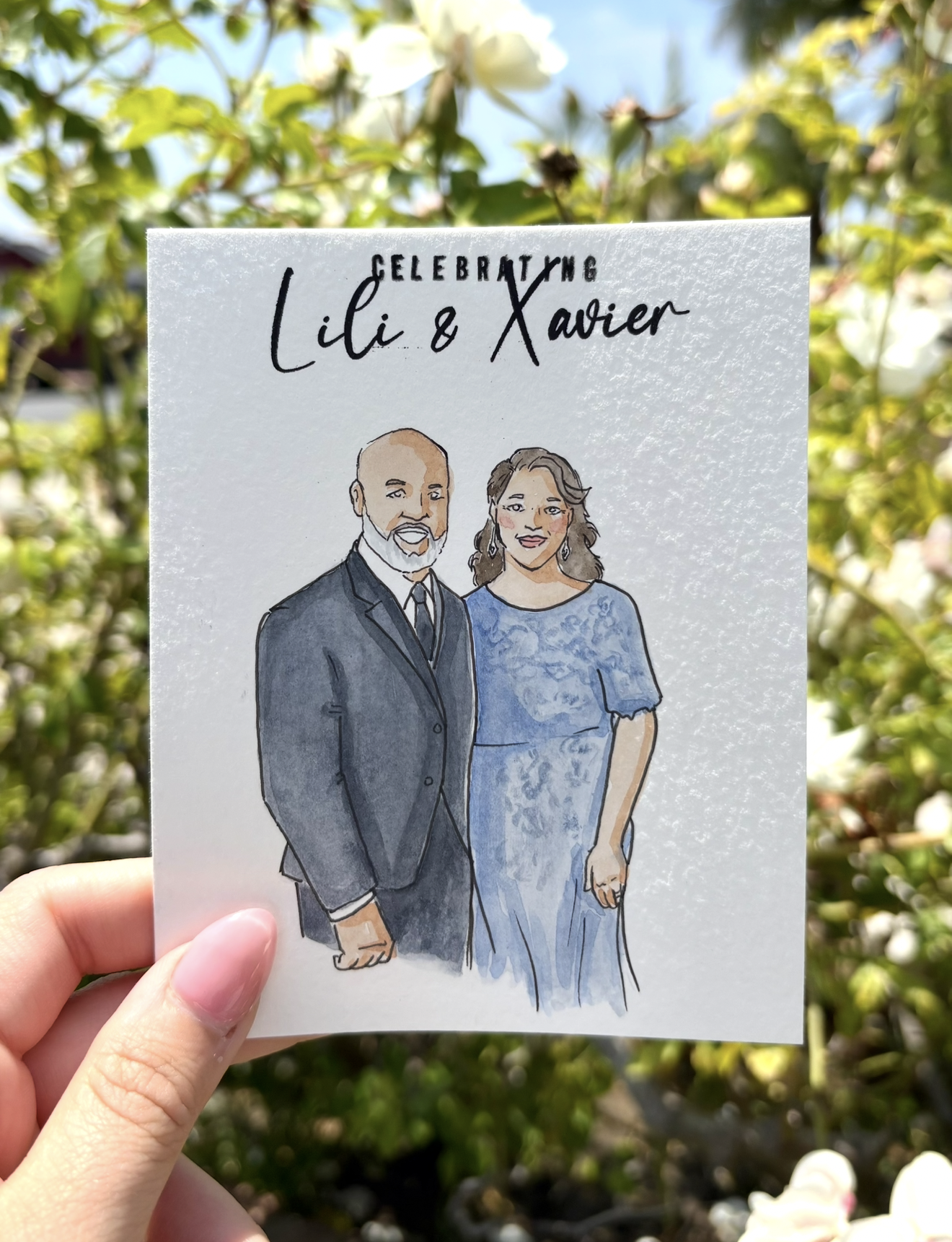 A hand holding a illustrated card with drawings of a man in a suit and a woman in a blue dress, against a background of flowering bushes.