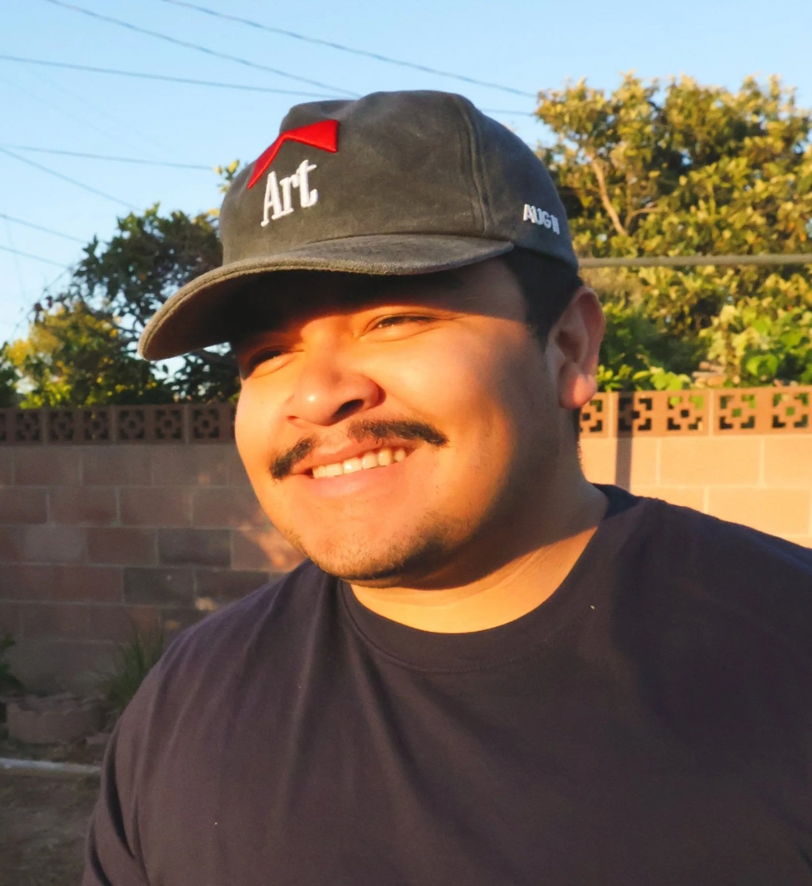 Smiling young man with a mustache and beard wearing a black t-shirt and a cap with the word 'Art' embroidered on it, standing outdoors in front of a brick wall and trees during sunset.