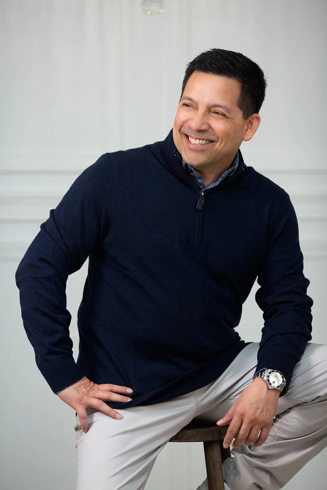 A smiling man sitting on a wooden stool, wearing a navy blue pullover and light-colored pants, with a white wall in the background.
