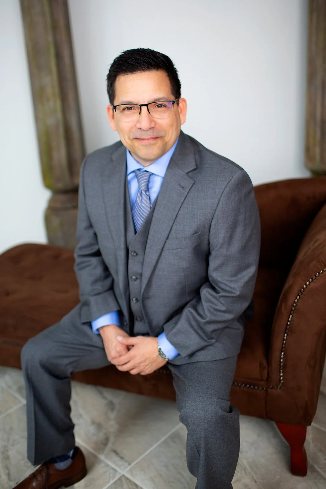 A man in a gray suit, blue shirt, and tie sitting on a brown leather sofa, wearing glasses, in a room with wooden pillars and light-colored walls.