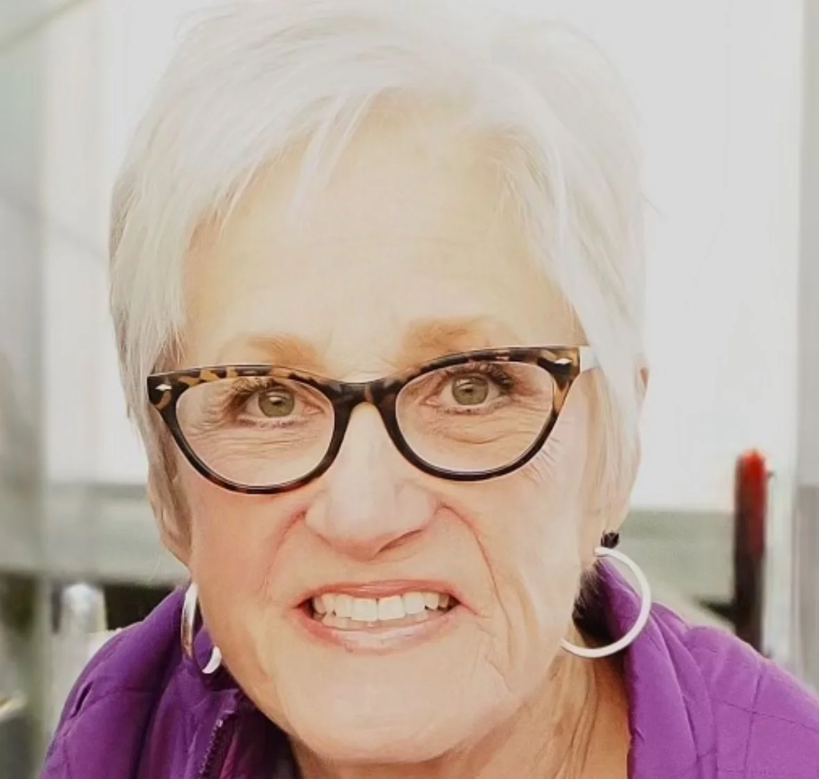 Close-up portrait of an elderly woman with short white hair, wearing tortoiseshell glasses, hoop earrings, and a purple jacket, smiling in an indoor setting.