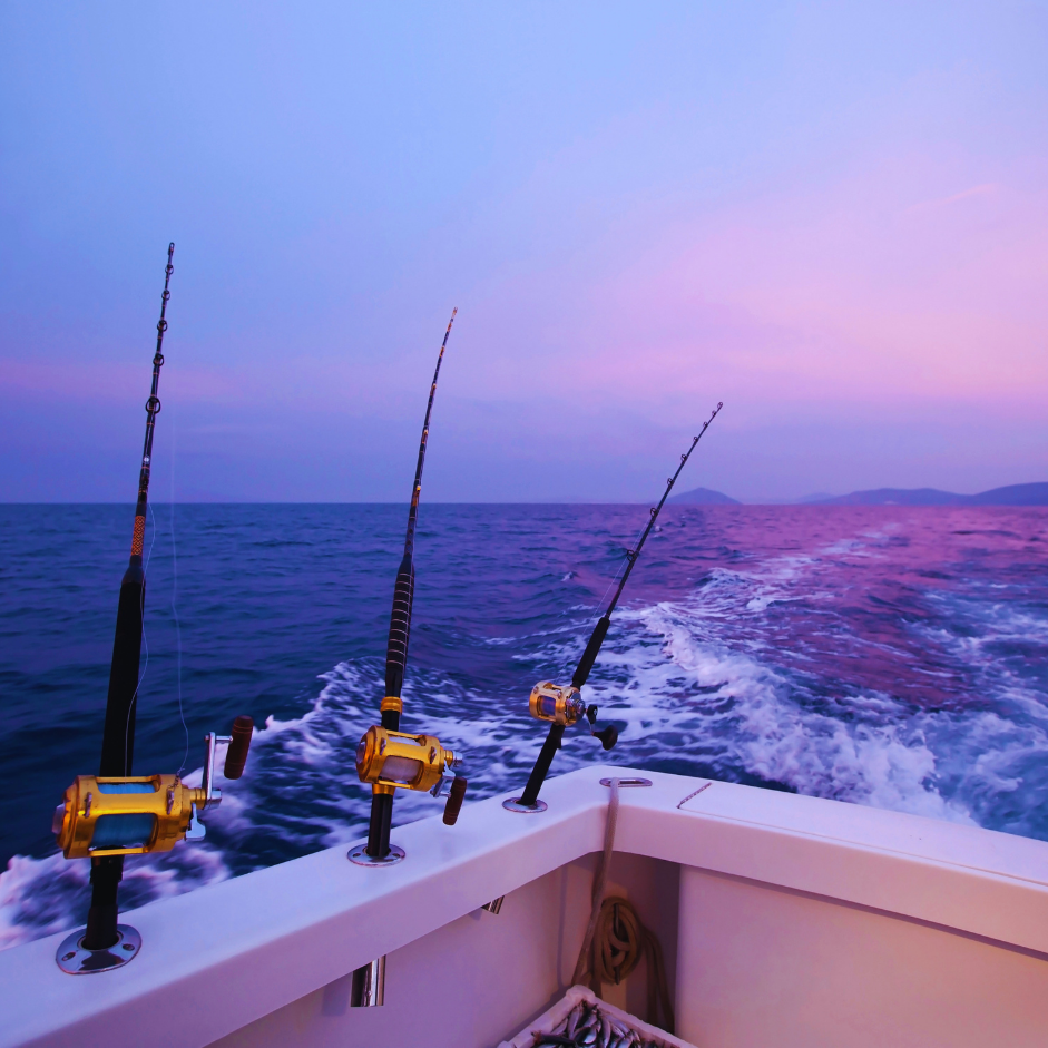 Three fishing rods mounted on the back of a boat as it moves through the ocean during sunset or sunrise, creating a wake in the water, with distant landforms on the horizon and a purple-pink sky.