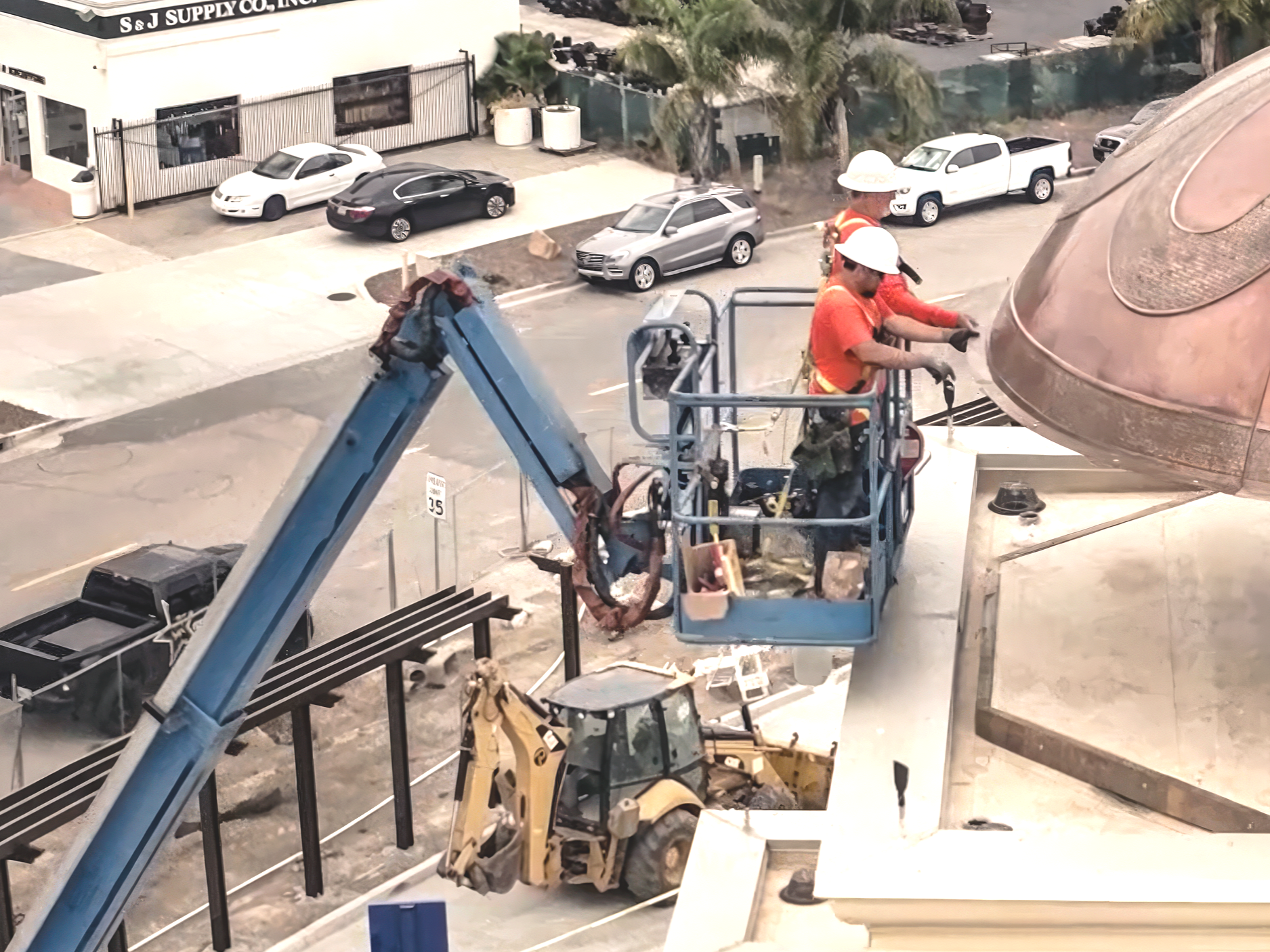 Construction workers in orange safety shirts and white helmets working on the exterior of a building, with several parked cars and trees in the background.